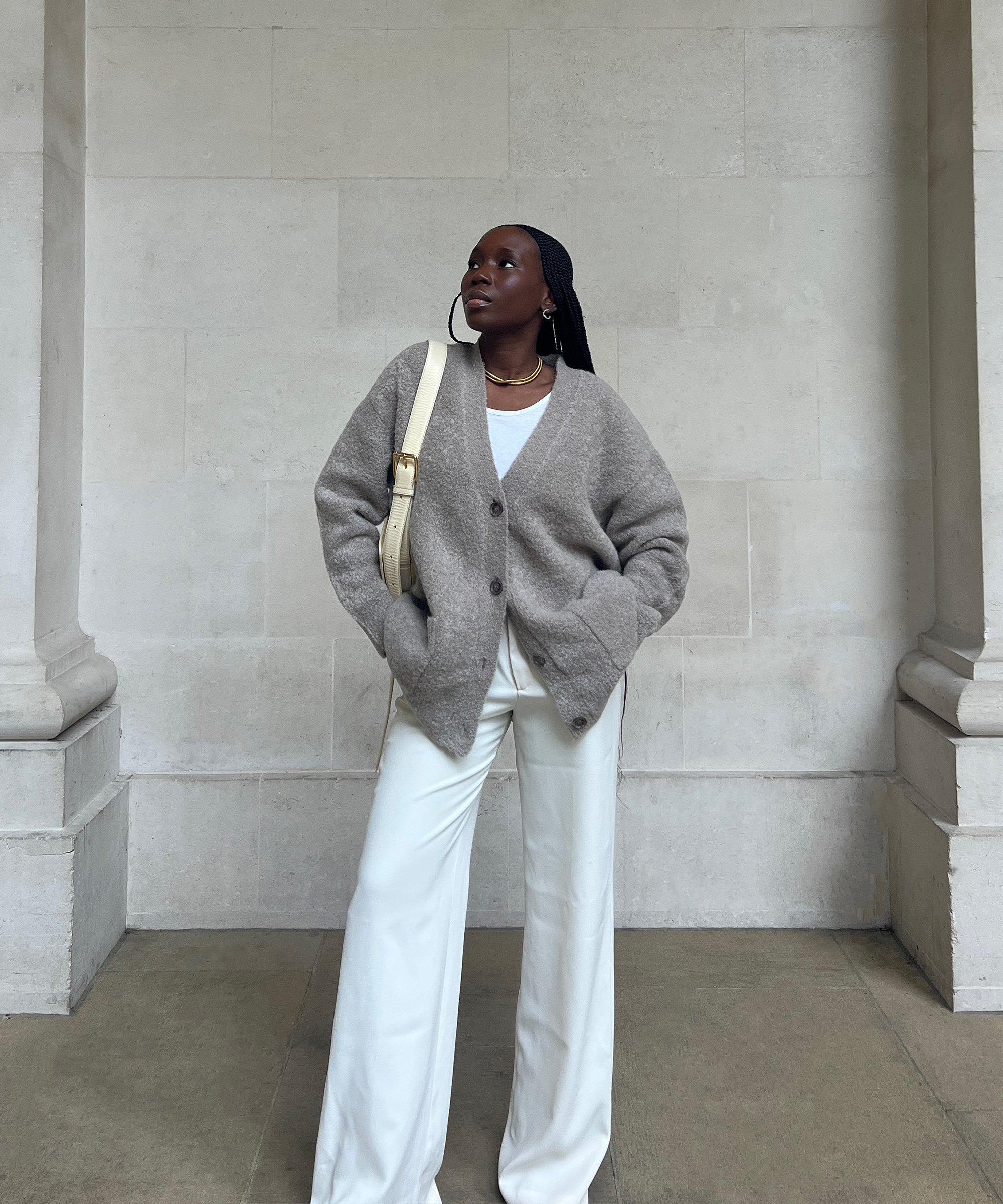 Danielle Jinadu stands confidently in a stone corridor, wearing a grey cardigan, white trousers, and a white top, with a cream shoulder bag. She looks slightly to the side with her hands in her pockets.