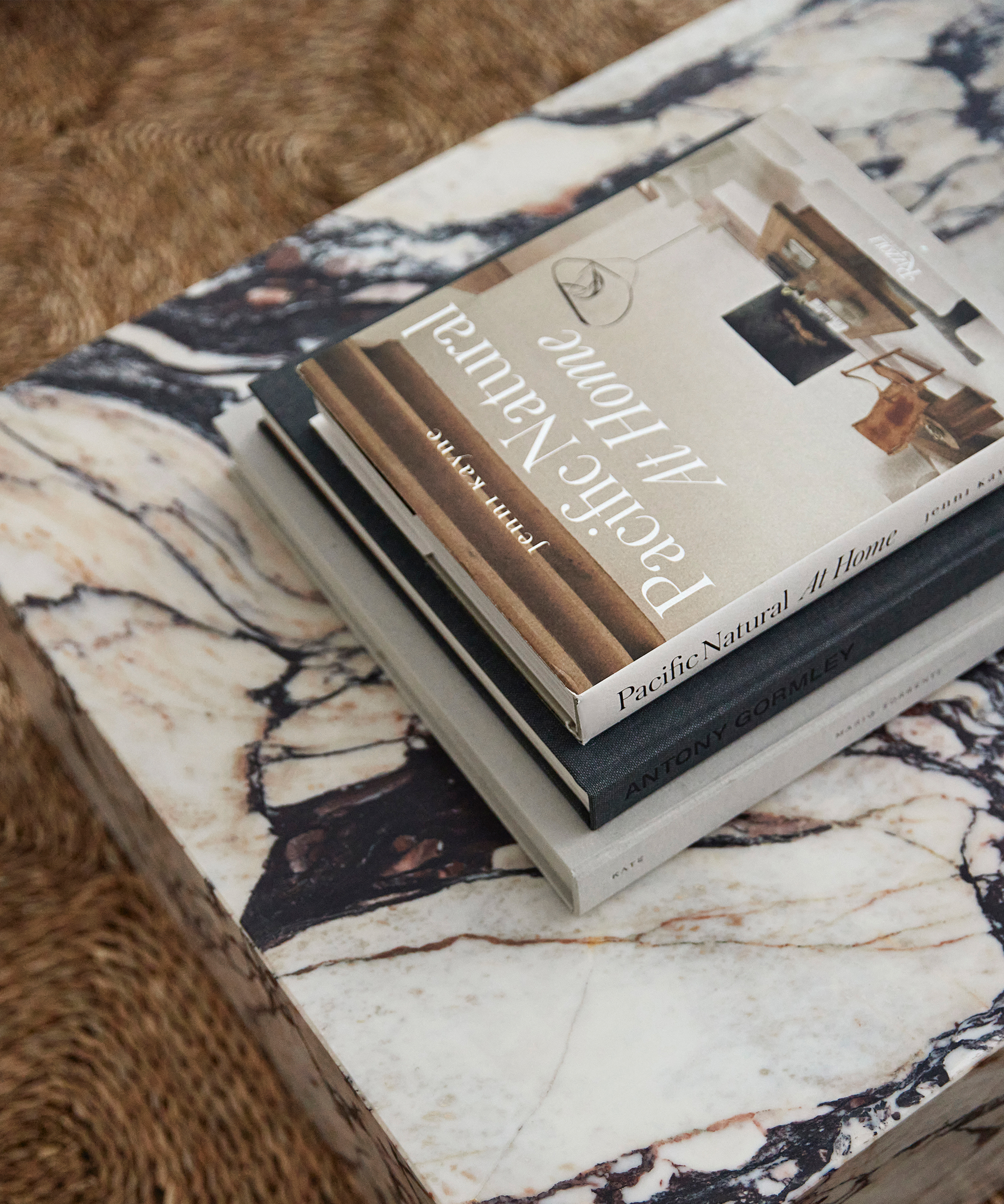 A stack of books, including Pacific Natural At Home and Antony Gormley, sits on a marble coffee table with striking veining in the living room of this Paige Pettit home. The textured, woven surface underneath adds inviting warmth.