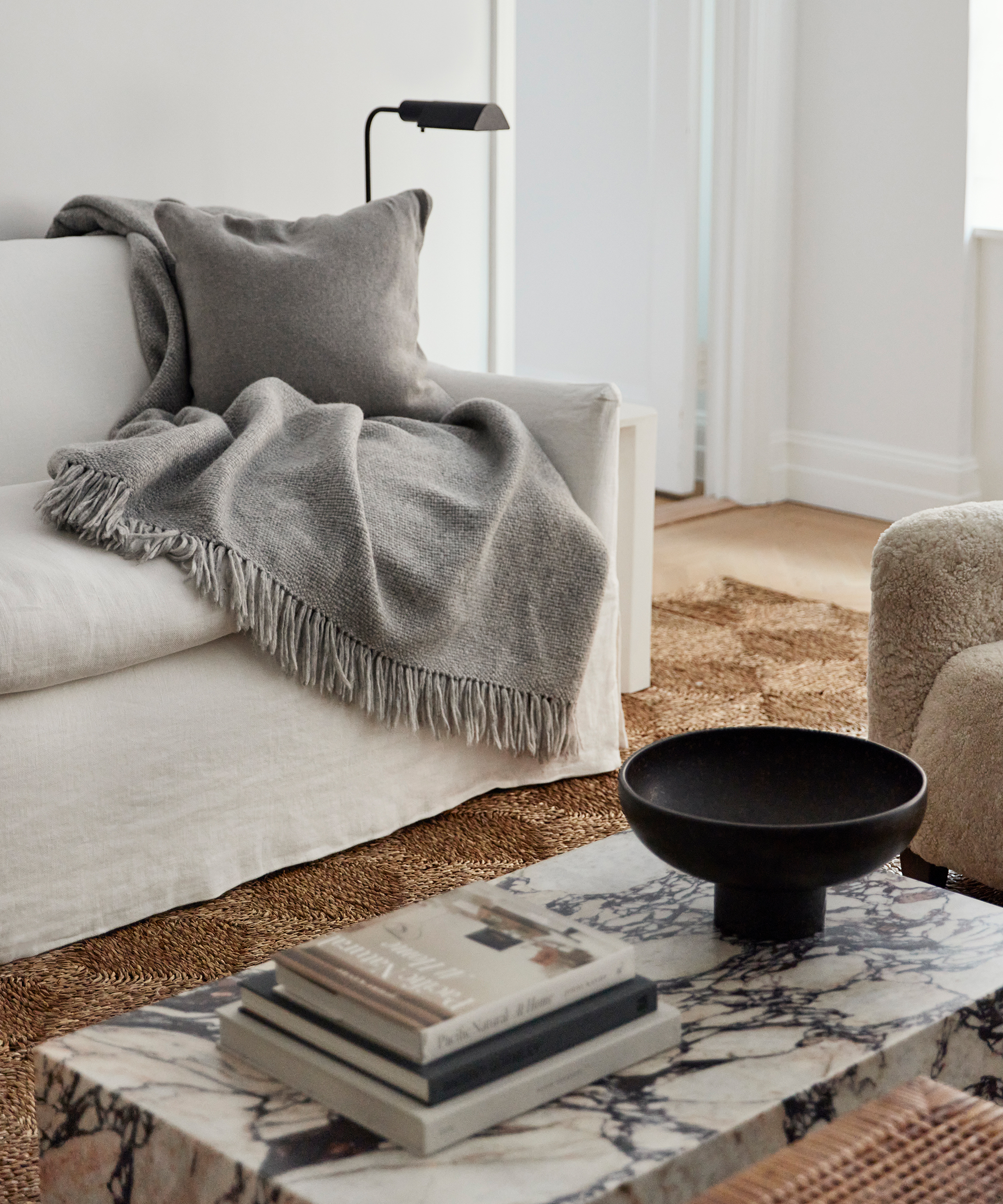 A cozy living room in true Paige Pettit Home style, featuring a white sofa with a gray throw blanket and pillow, a marble coffee table with books and a black bowl, a textured rug, and a minimal black lamp.