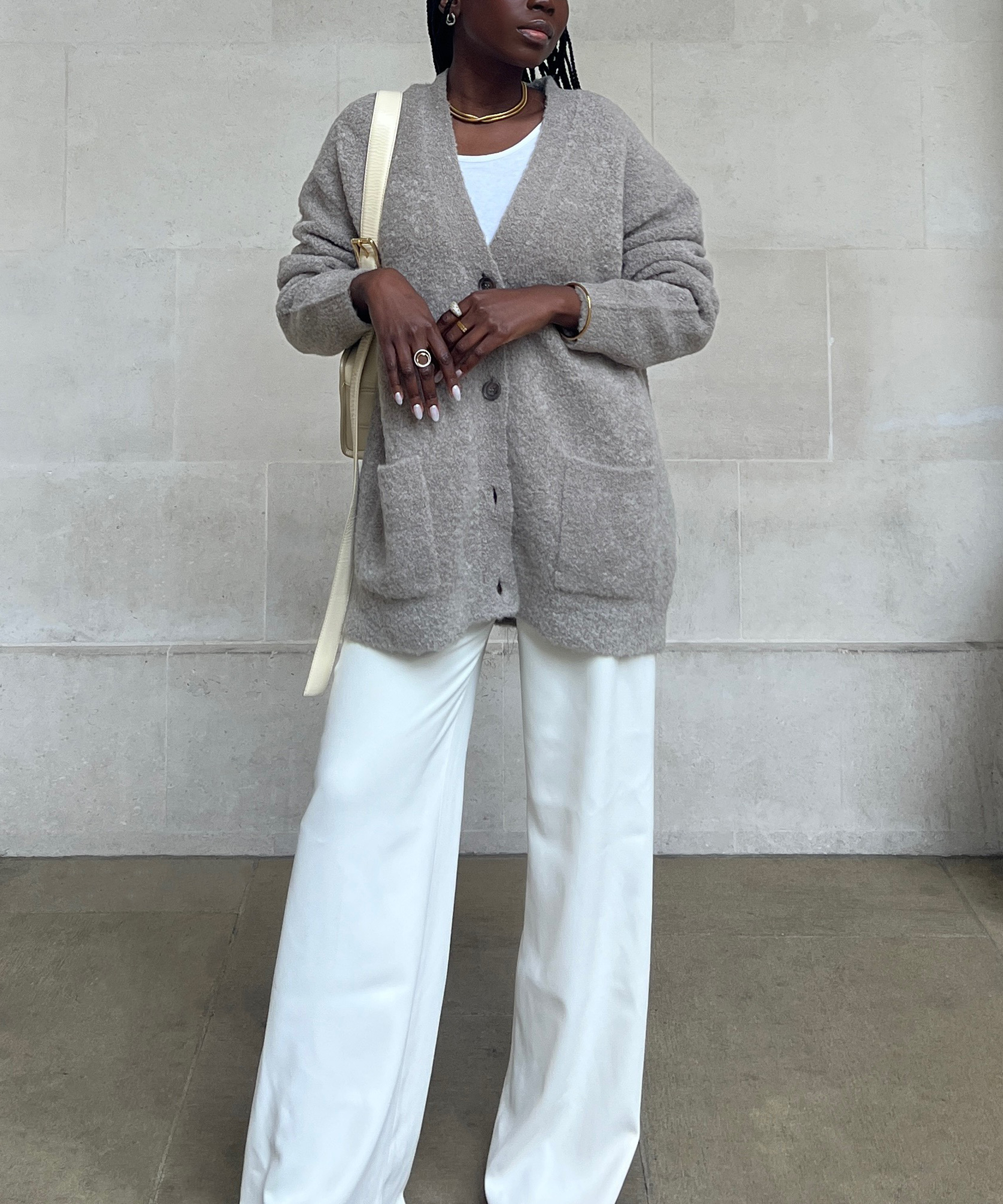 Danielle Jinadu wears a beige cardigan over a white top with wide-leg white pants, accessorized with gold jewelry and a cream bag, as she stands against a neutral stone wall.