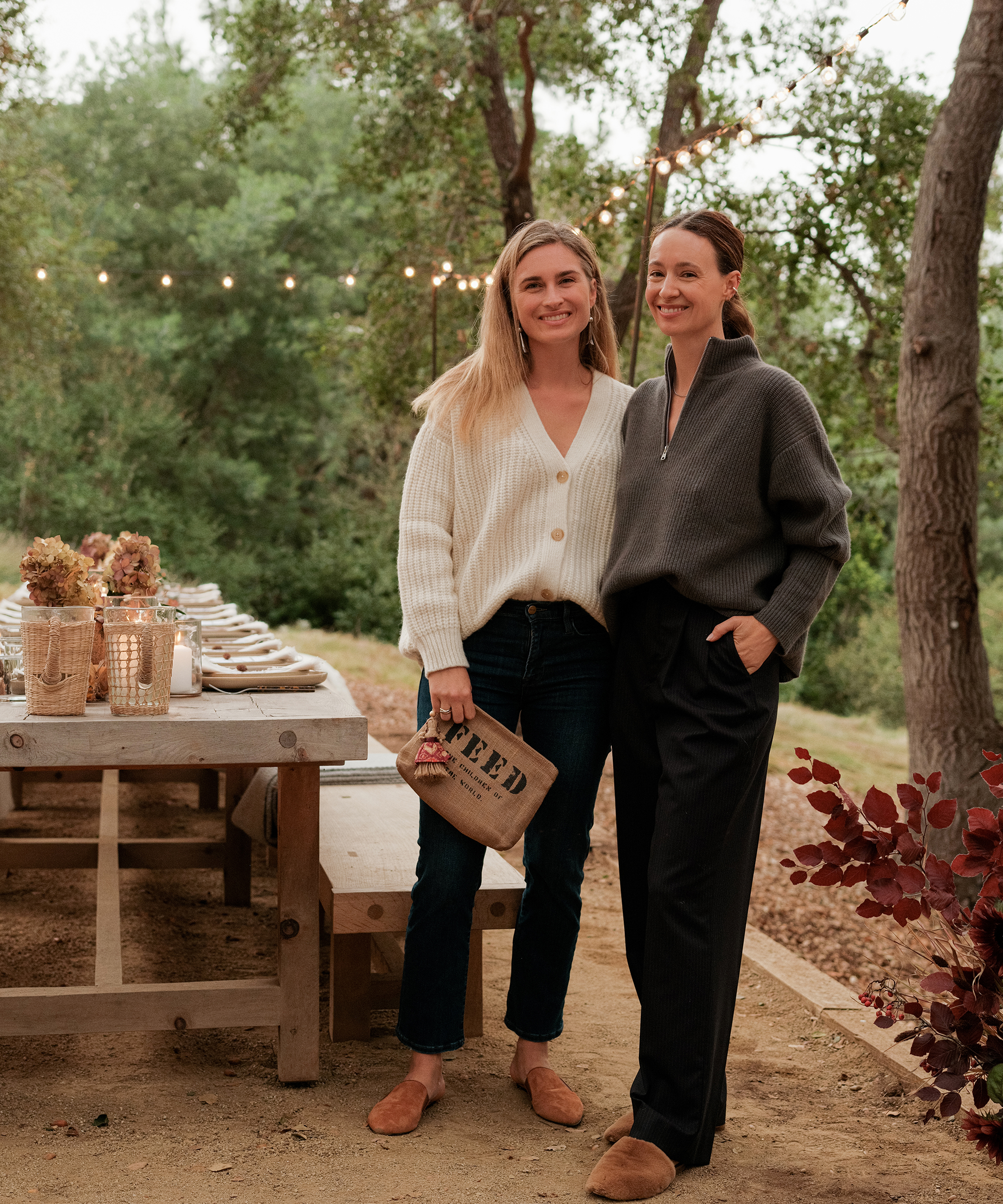Two women stand smiling outdoors beside a wooden table set for a meal, with FEED pouch in hand, string lights above, and trees behind. Both wear cozy sweaters and flat shoes, creating a warm and inviting scene.