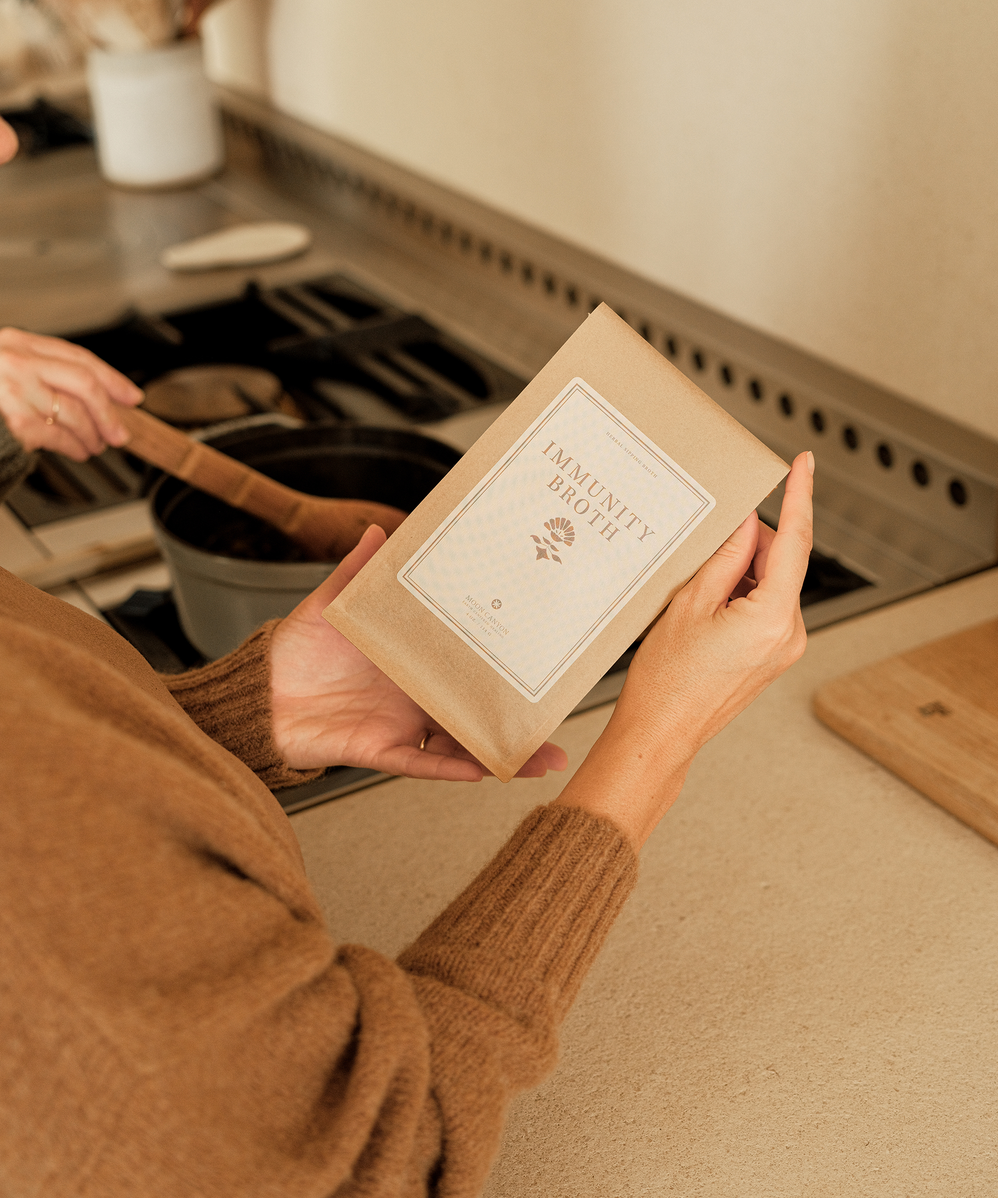 A person in a brown sweater holds a packet labeled Immunity Broth from Moon Canyon Healing while stirring a pot on a stovetop in a cozy kitchen.