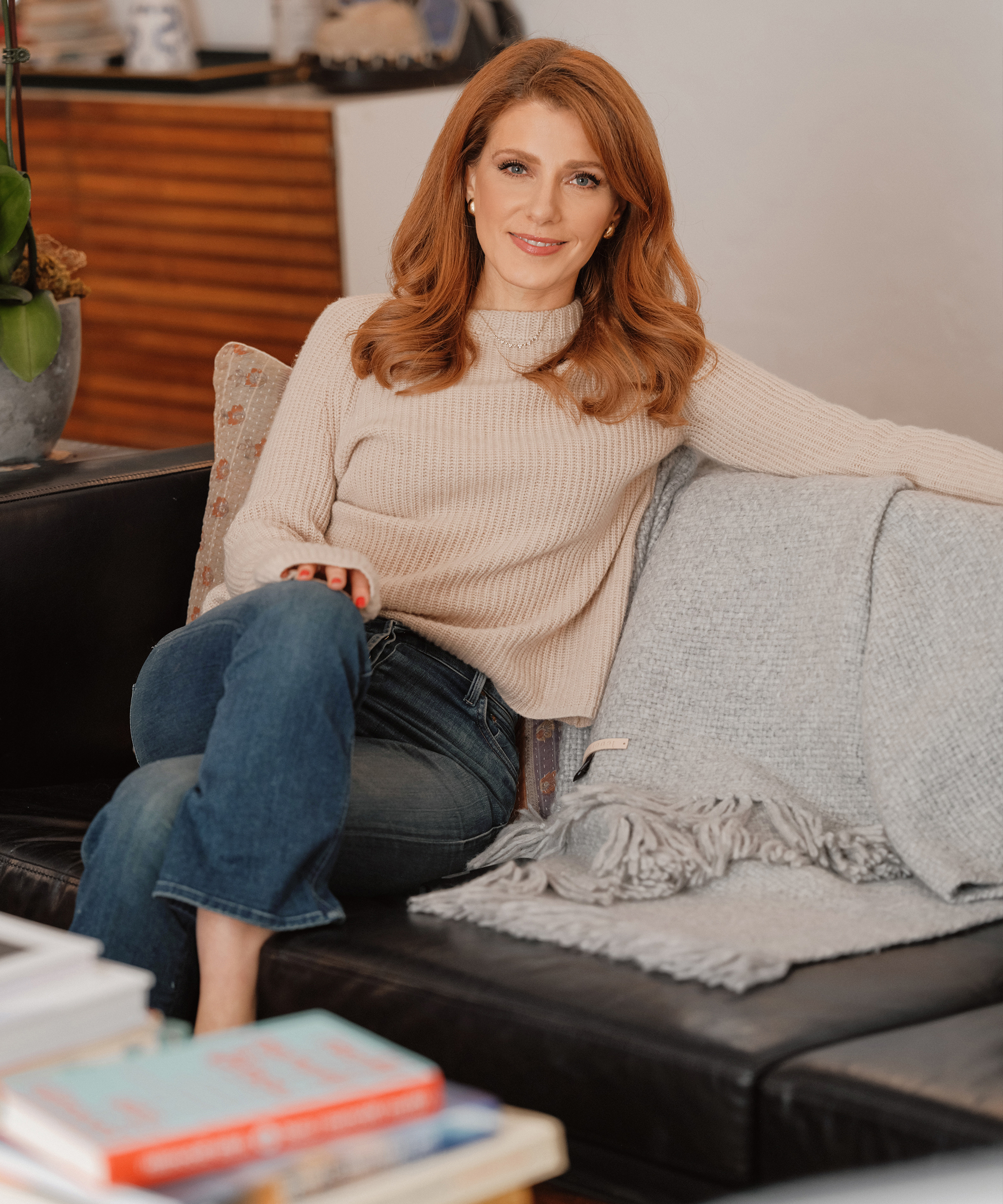 Julia Boorstin, with her long red hair, sits comfortably on a dark sofa in a cream sweater and blue jeans, smiling at the camera. A light gray throw blanket drapes beside her, and books are stacked on the table in the foreground.