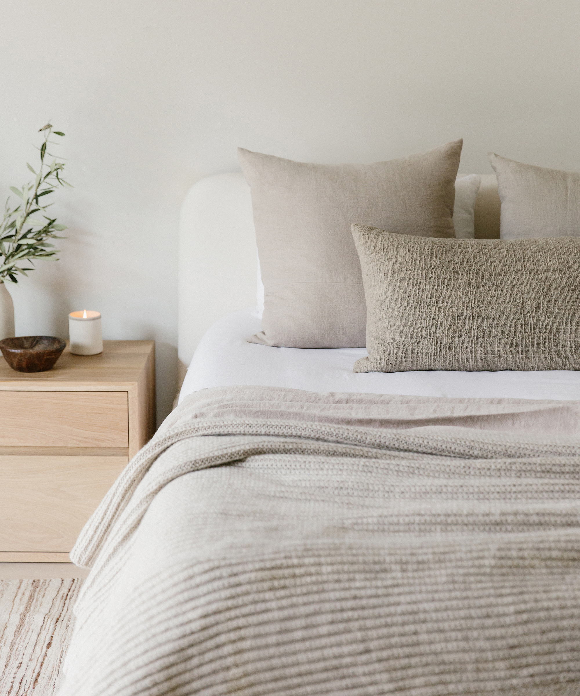 A neatly made bed with textured neutral bedding sits beside a light wooden nightstand holding a lit candle, bowl, and vase with leafy greenery. The overall decor is minimalist and calming—perfect inspiration for anyone seeking simple interior design tips.
