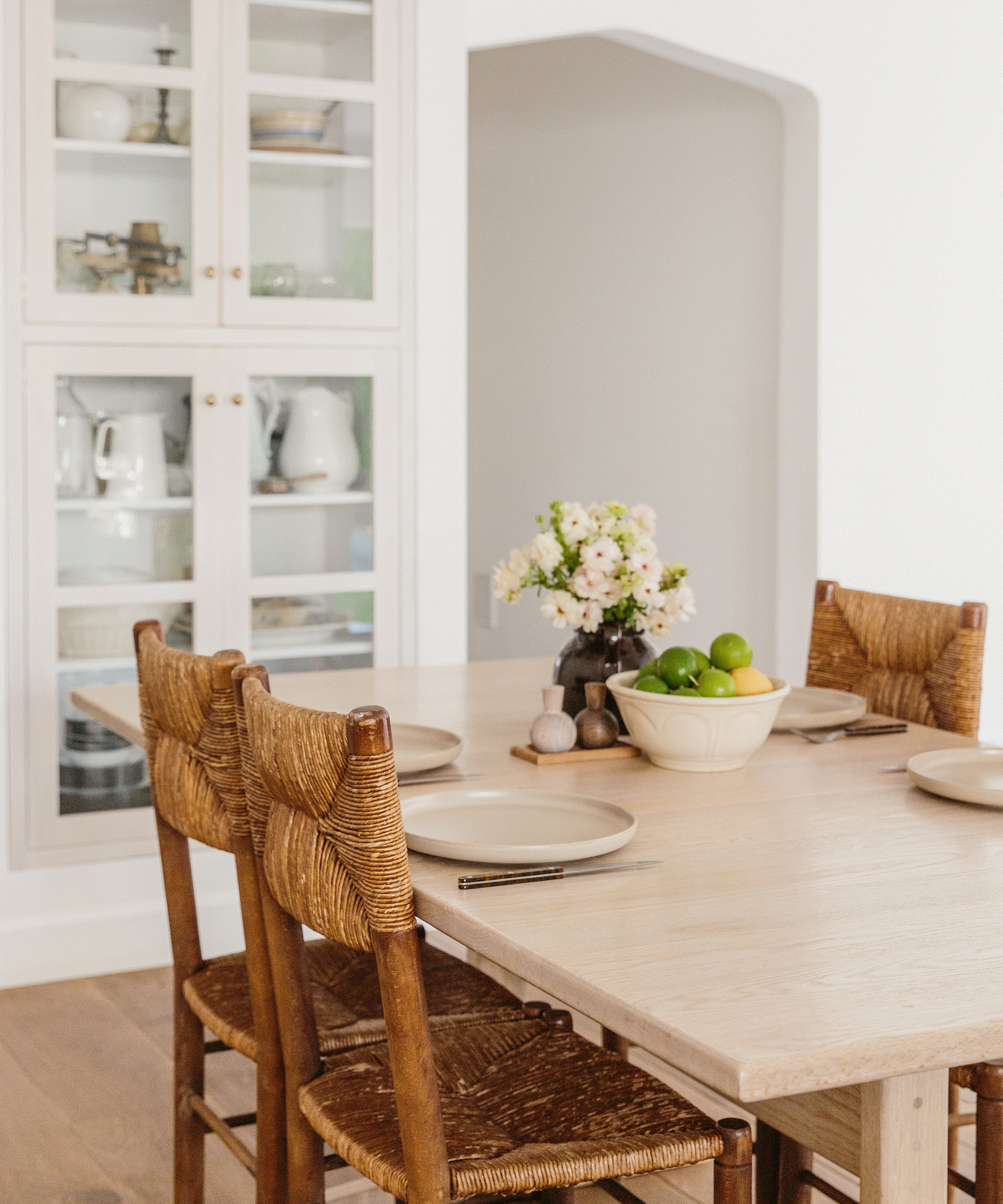 A light wooden dining table with four woven chairs, set with plates and a bowl of fresh fruit, channels California design. A vase of flowers sits at the center, while a glass-fronted cabinet with dishes decorates the background.