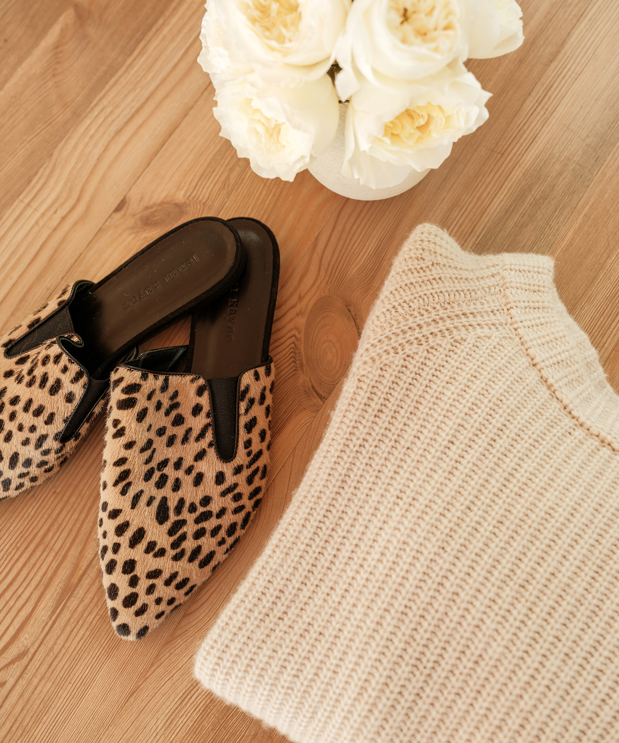 folded oatmeal cashmere sweater next to cheetah mule slides and white flowers on hardwood floors