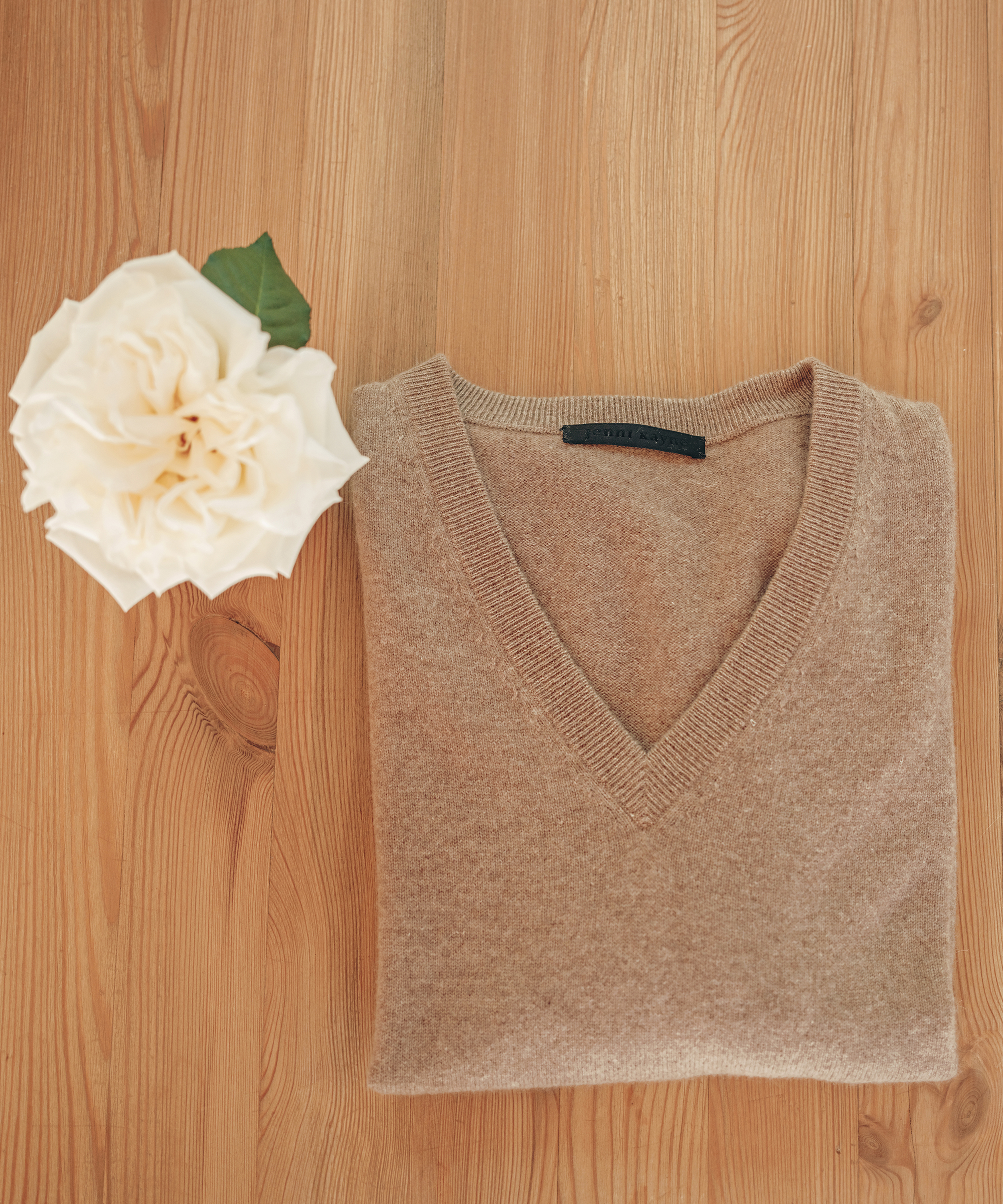 folded cashmere v-neck sweater in camel on hardwood floor with a white flower