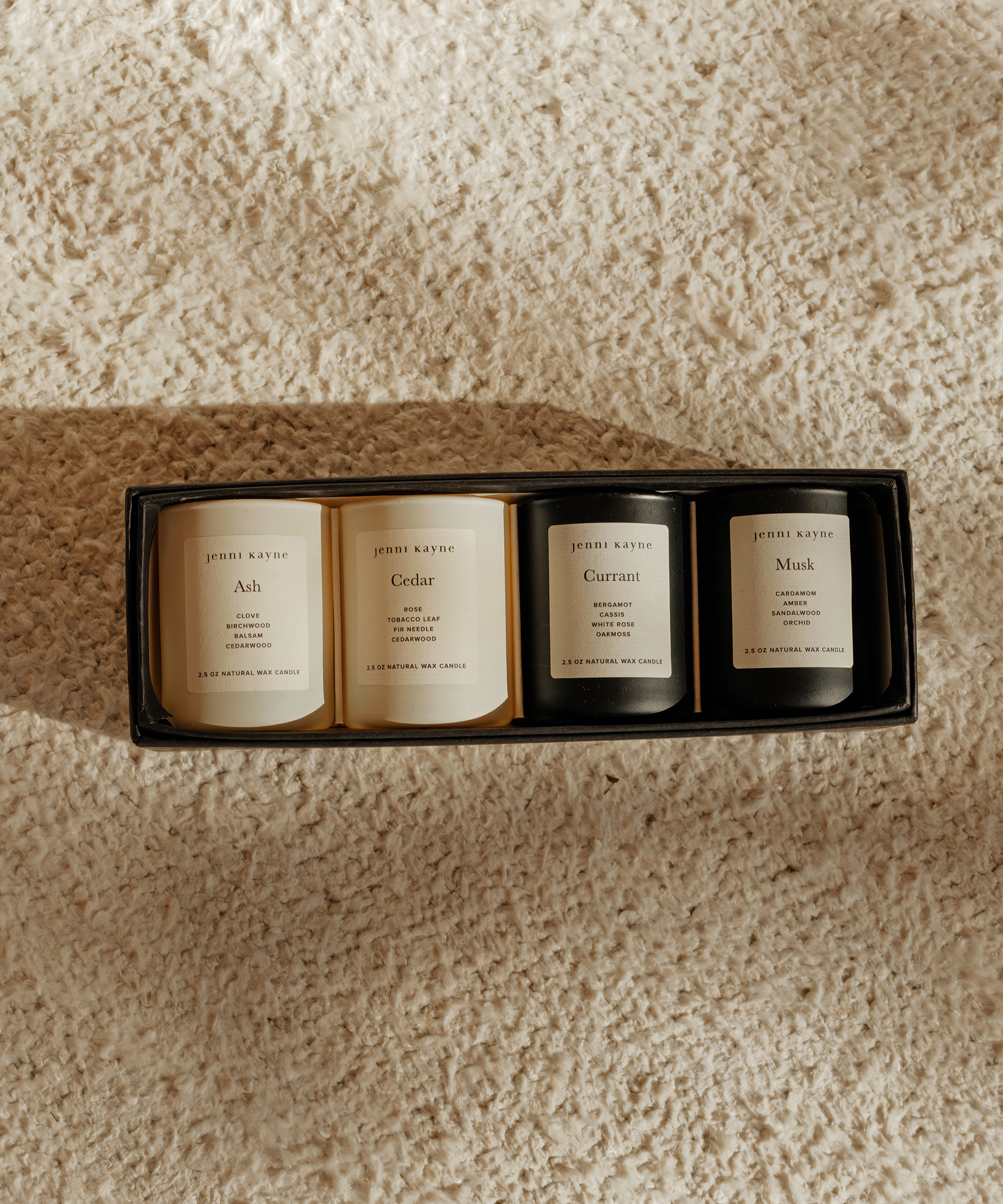 Four Jennie Kayne candles labeled Ash, Cedar, Currant, and Musk are arranged in a box on a textured beige surface, lit by warm natural light—perfect for thoughtful Valentine’s Day gift ideas for him.