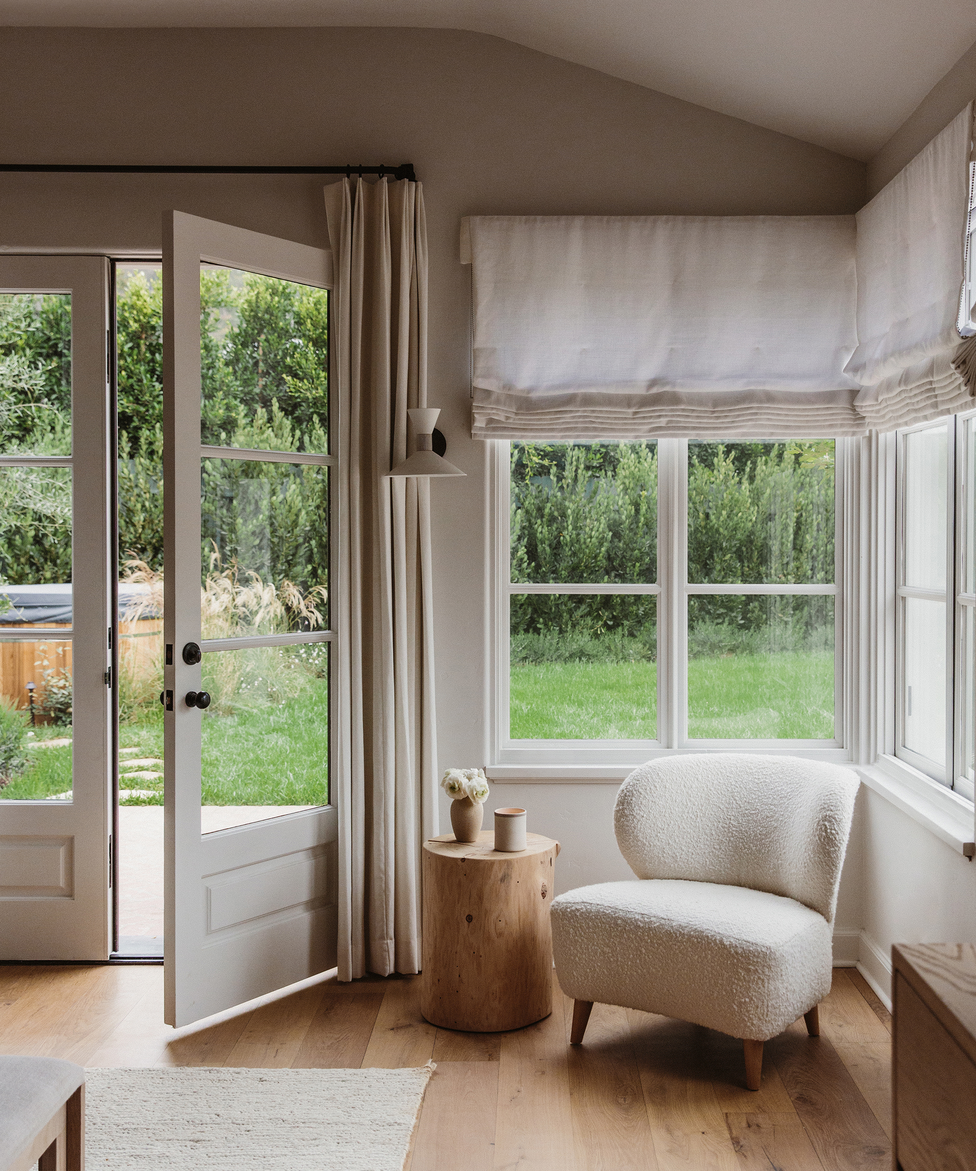 corner of bedroom with ivory boucle accent chair under a window next to a cedar side table with door open to outside