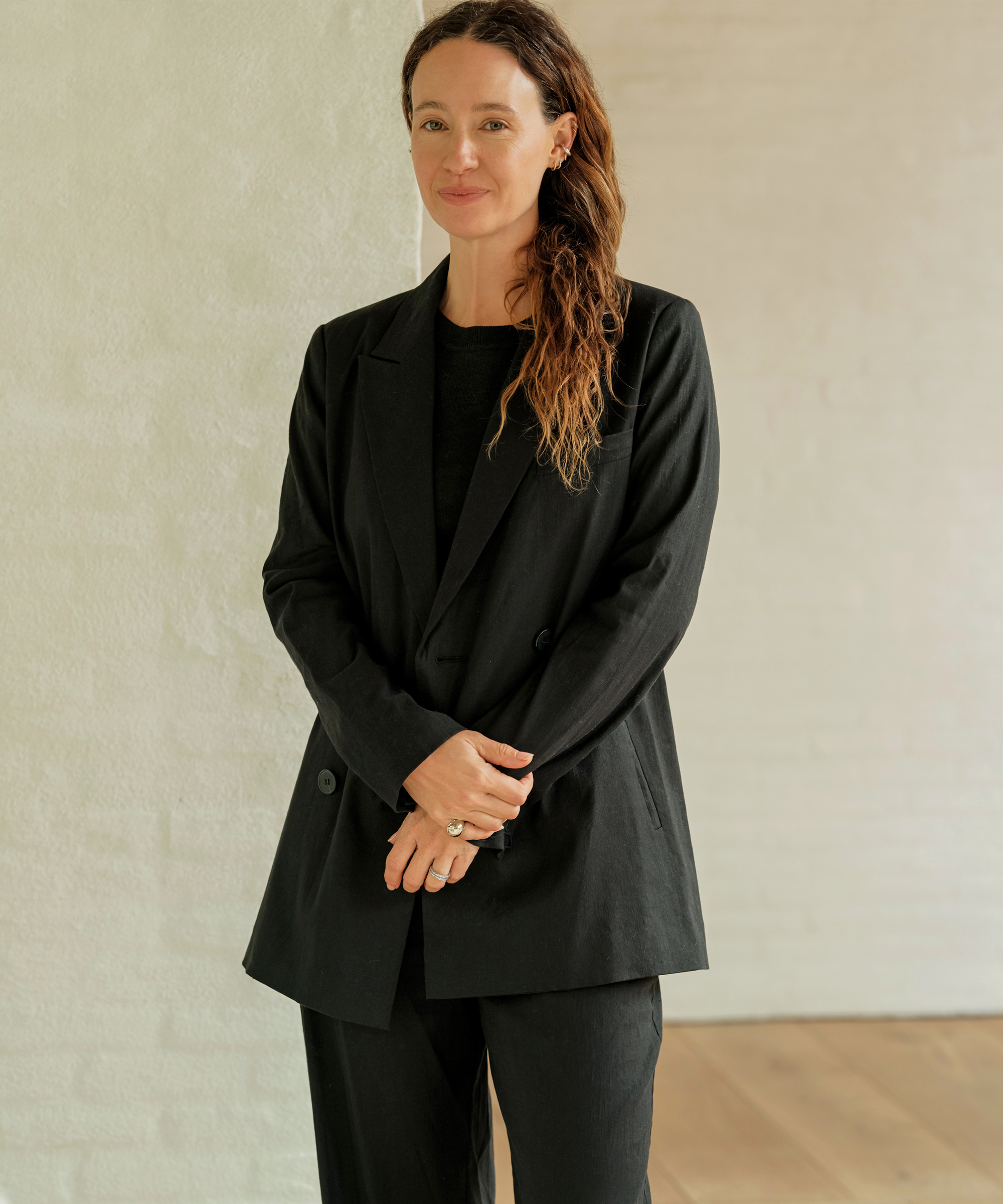A woman with wavy brown hair wearing a chic black suit stands indoors against a light-colored wall, smiling softly with her hands clasped in front—a perfect touch of spring style elegance.