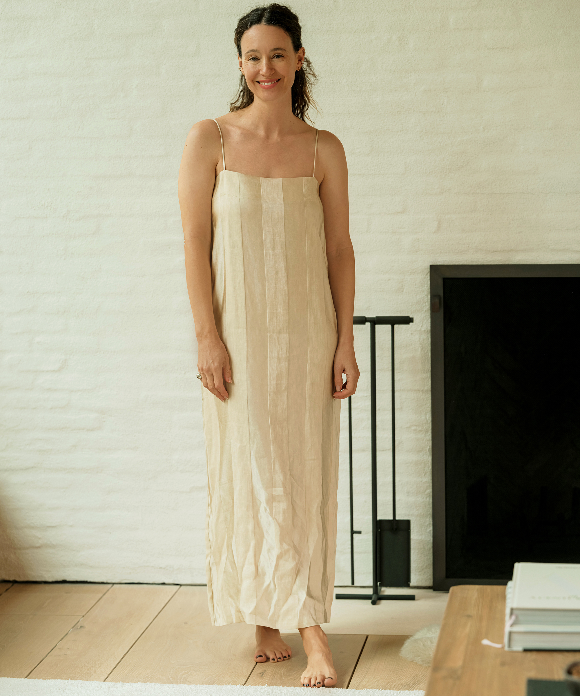 A woman with long dark hair stands barefoot indoors, wearing a loose, light-colored, sleeveless dress that reflects an effortless spring style. She is smiling softly, with a white brick wall and dark fireplace in the background.