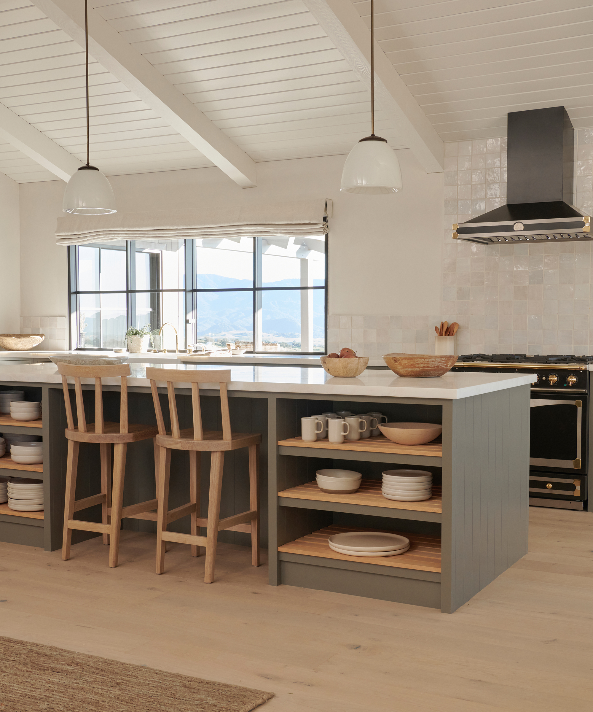 Bright, modern kitchen remodel featuring light wood floors, a large island with shelves stocked with dishes, two wooden chairs, pendant lights, a range with a black hood, and large windows letting in natural light.
