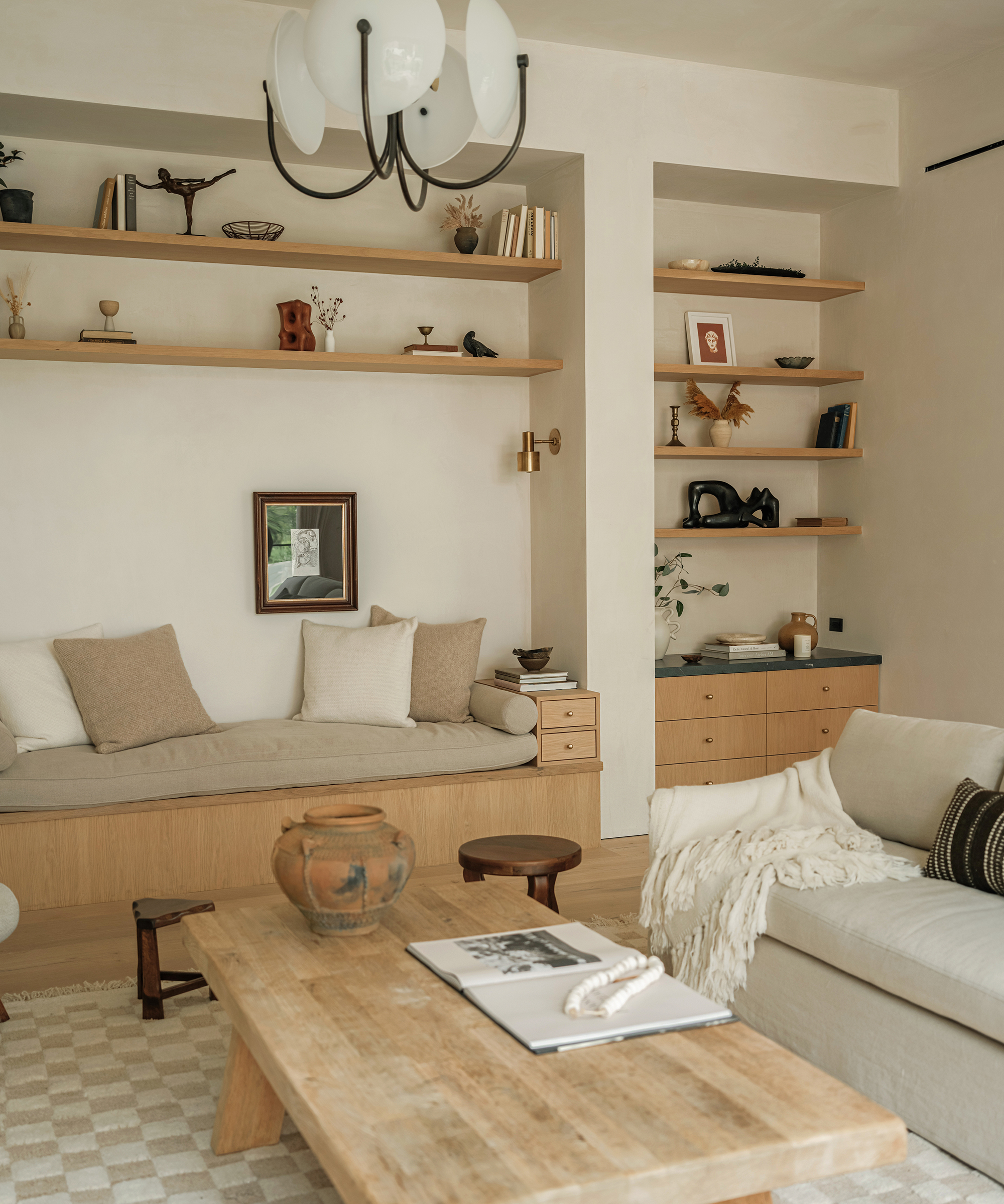 A cozy, modern studio city home living room with neutral tones, light wood furniture, open shelves displaying books and decor, a built-in bench with cushions, and a beige sofa with a throw blanket beside a wooden coffee table.