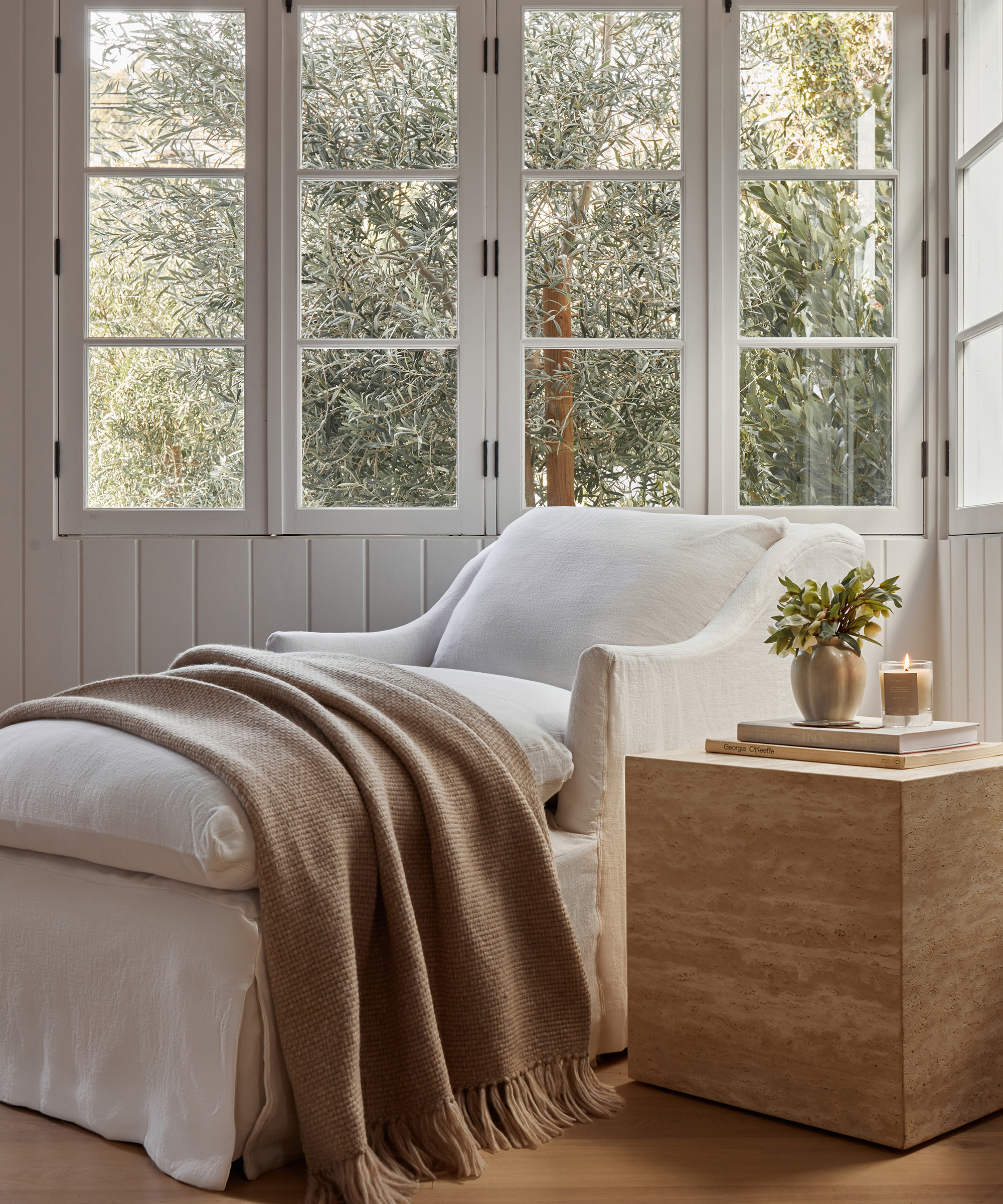 white linen chase in a sunlit corner with an oatmeal throw draped across next to a travertine side table