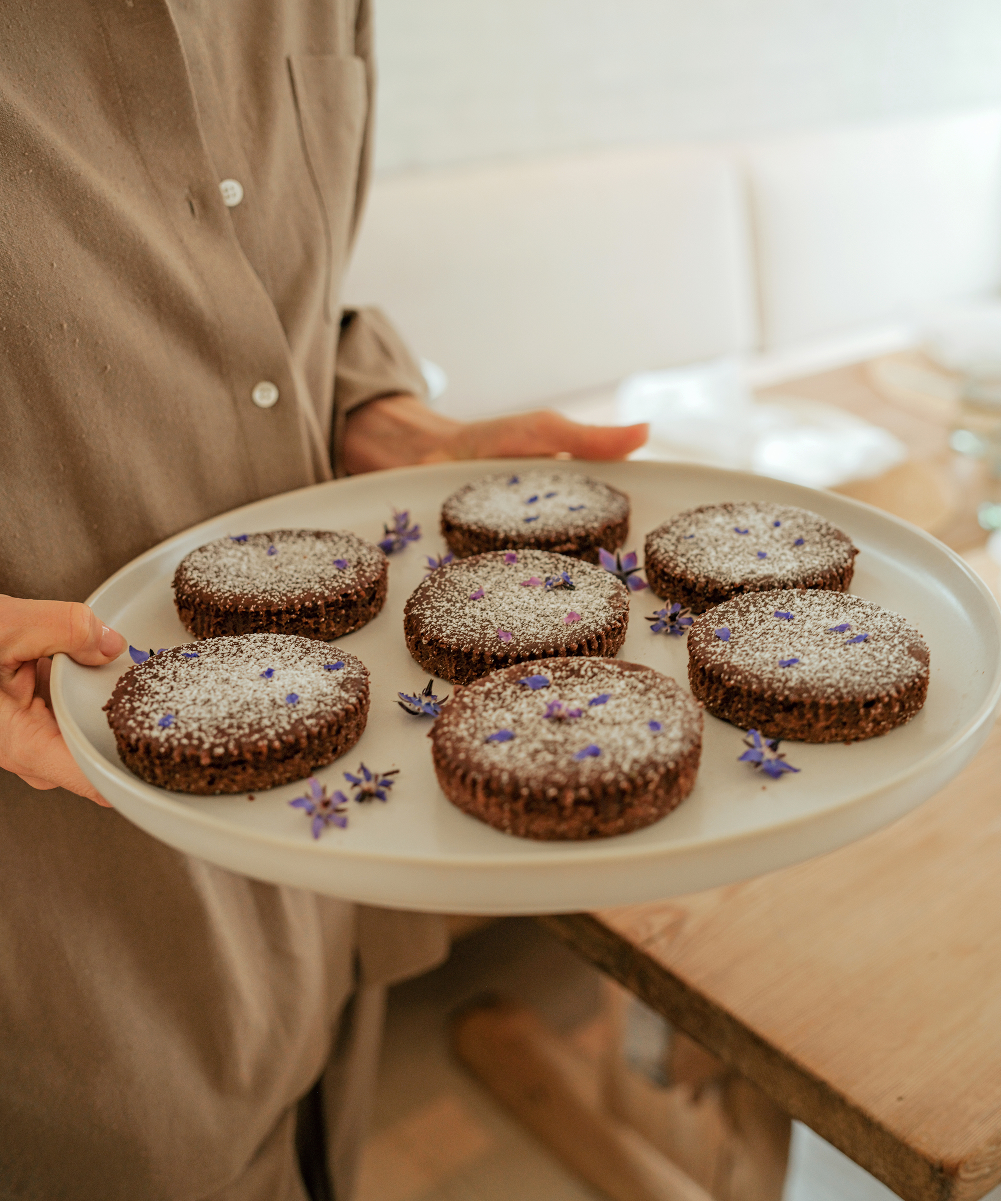 A person in a brown shirt holds a white plate with seven chocolate desserts topped with powdered sugar and small purple edible flowers. The gluten free quiche-inspired treats stand out against the softly blurred background.