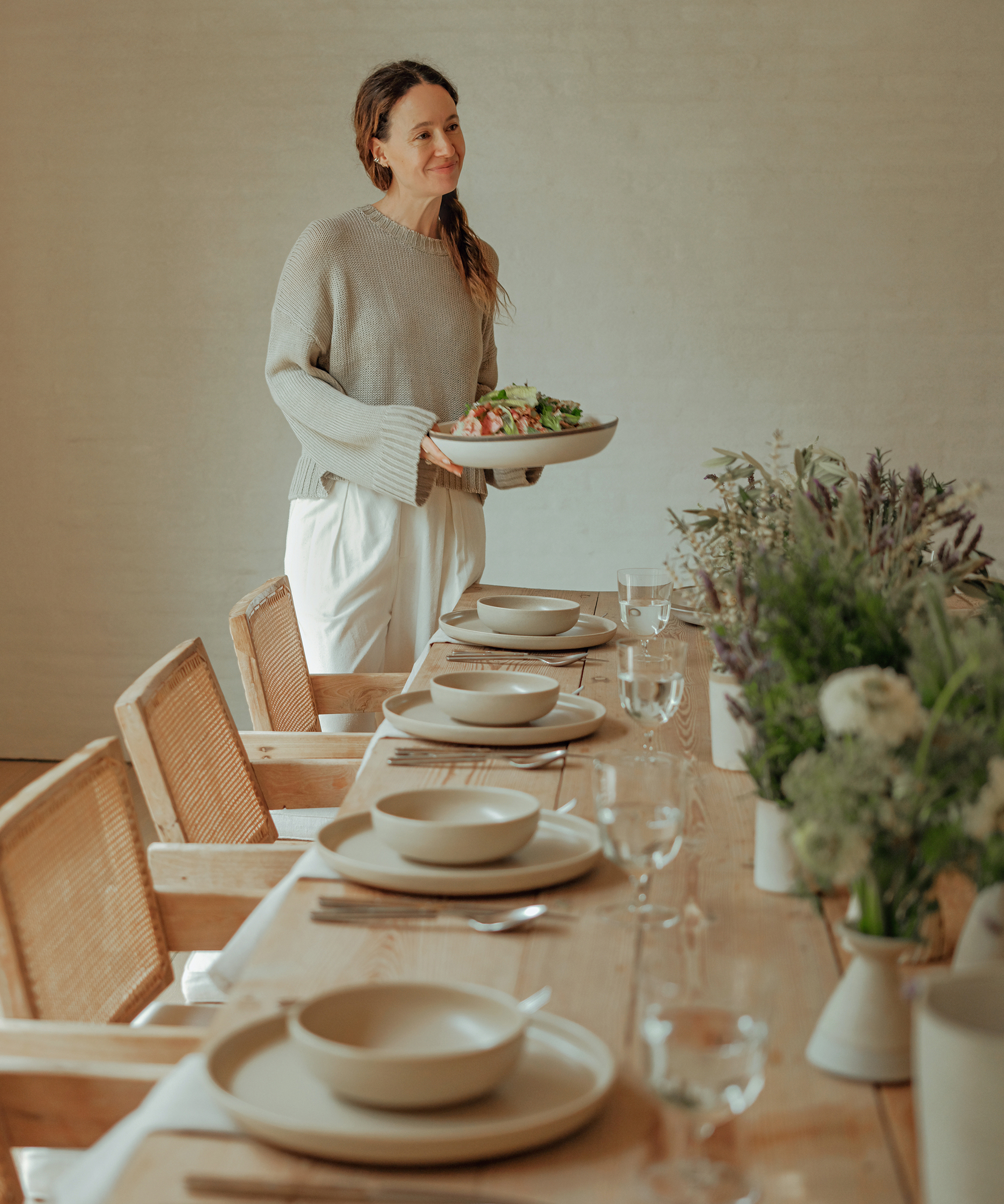 A woman in a light sweater and white pants holds a platter of food while standing next to a wooden dining table set with plates, bowls, glasses, and floral centerpieces.