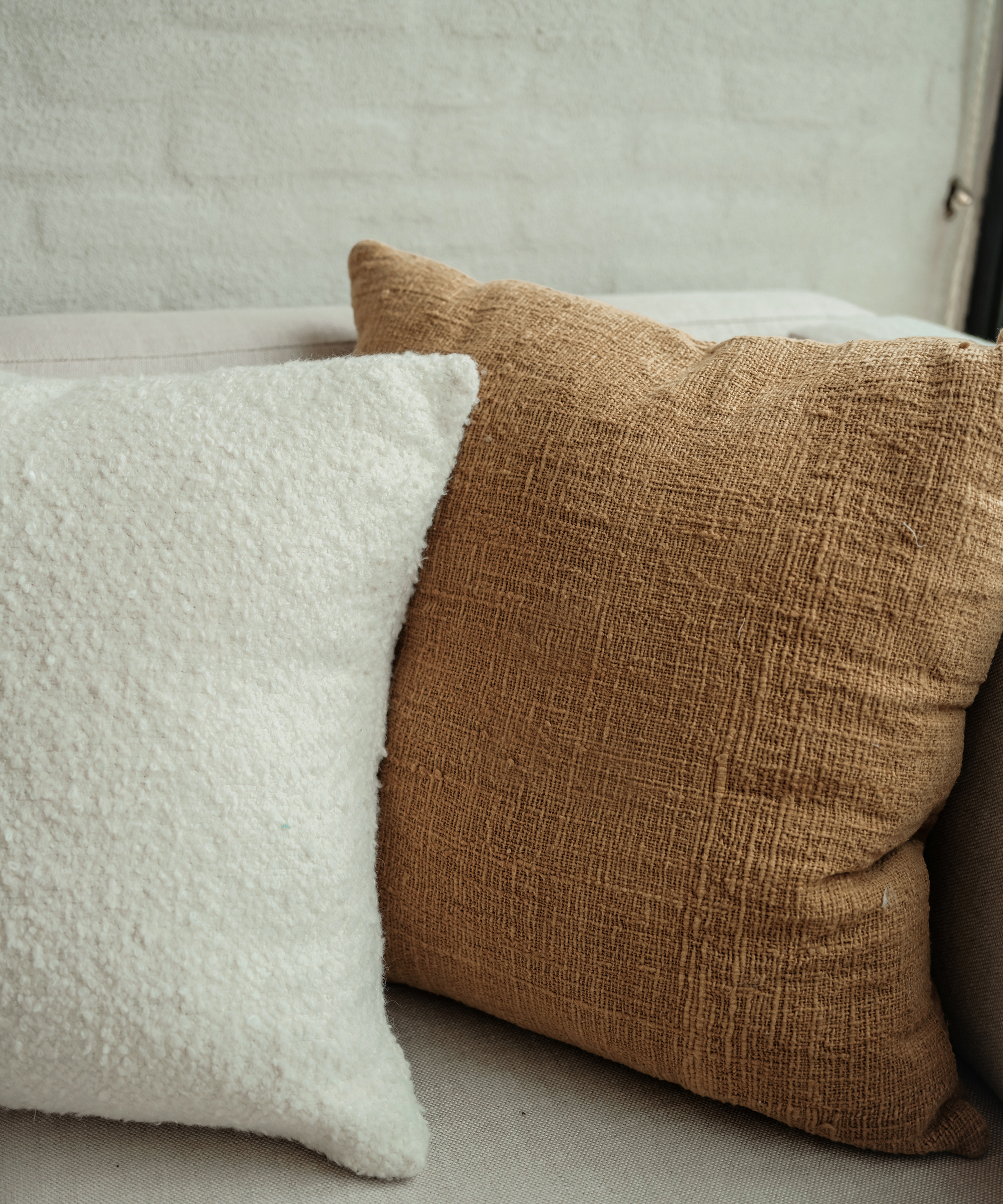Two pillows rest on a light-colored couch: one is white and fluffy, the other brown with textured fabric. This cozy scene creates a calming space—perfect for balancing hormones and unwinding against a white brick wall backdrop.