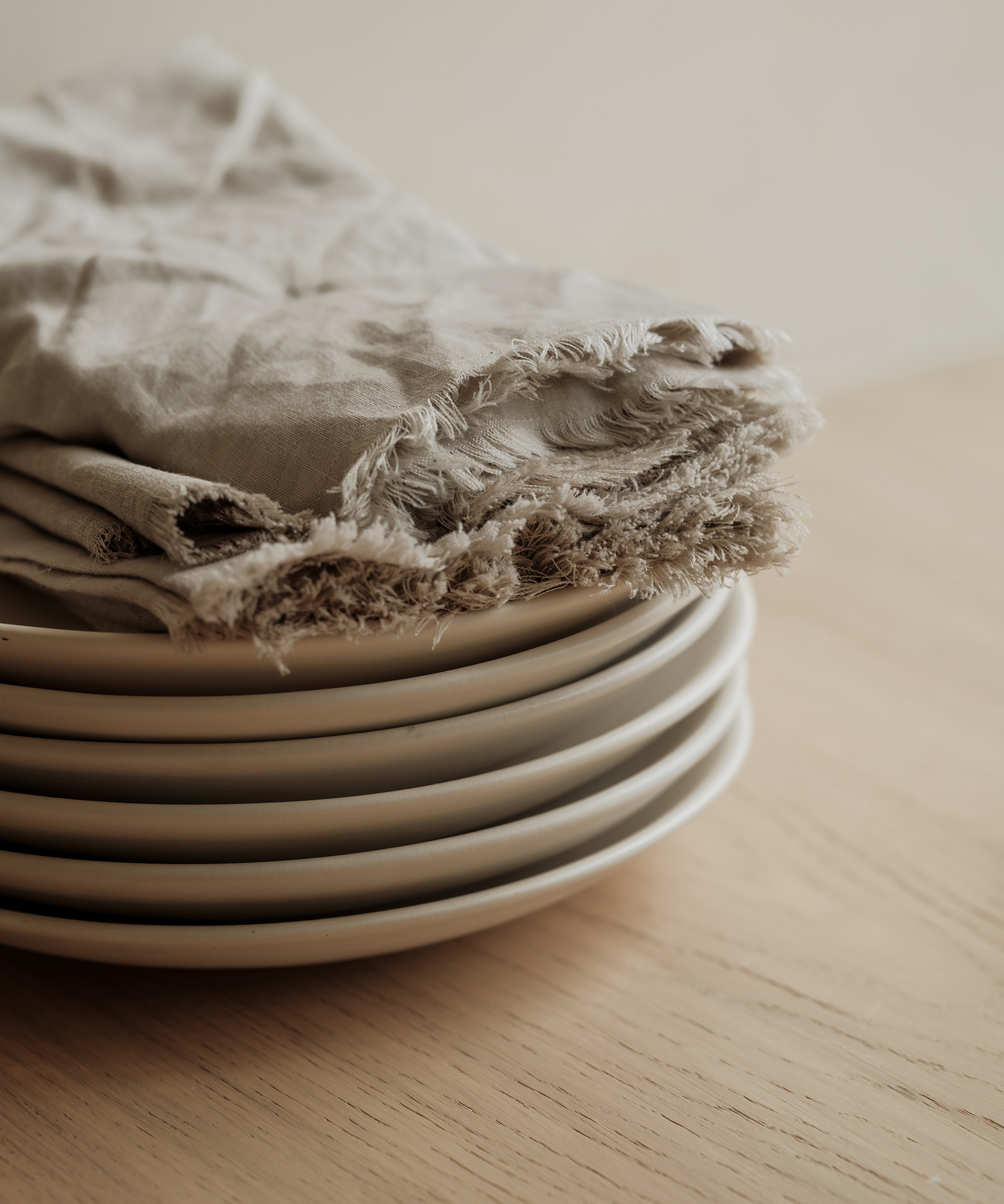A stack of white ceramic plates sits on a light wooden surface, topped with beige, frayed-edge linen napkins—perfect for serving your favorite cake recipes in this softly lit, minimalistic scene.