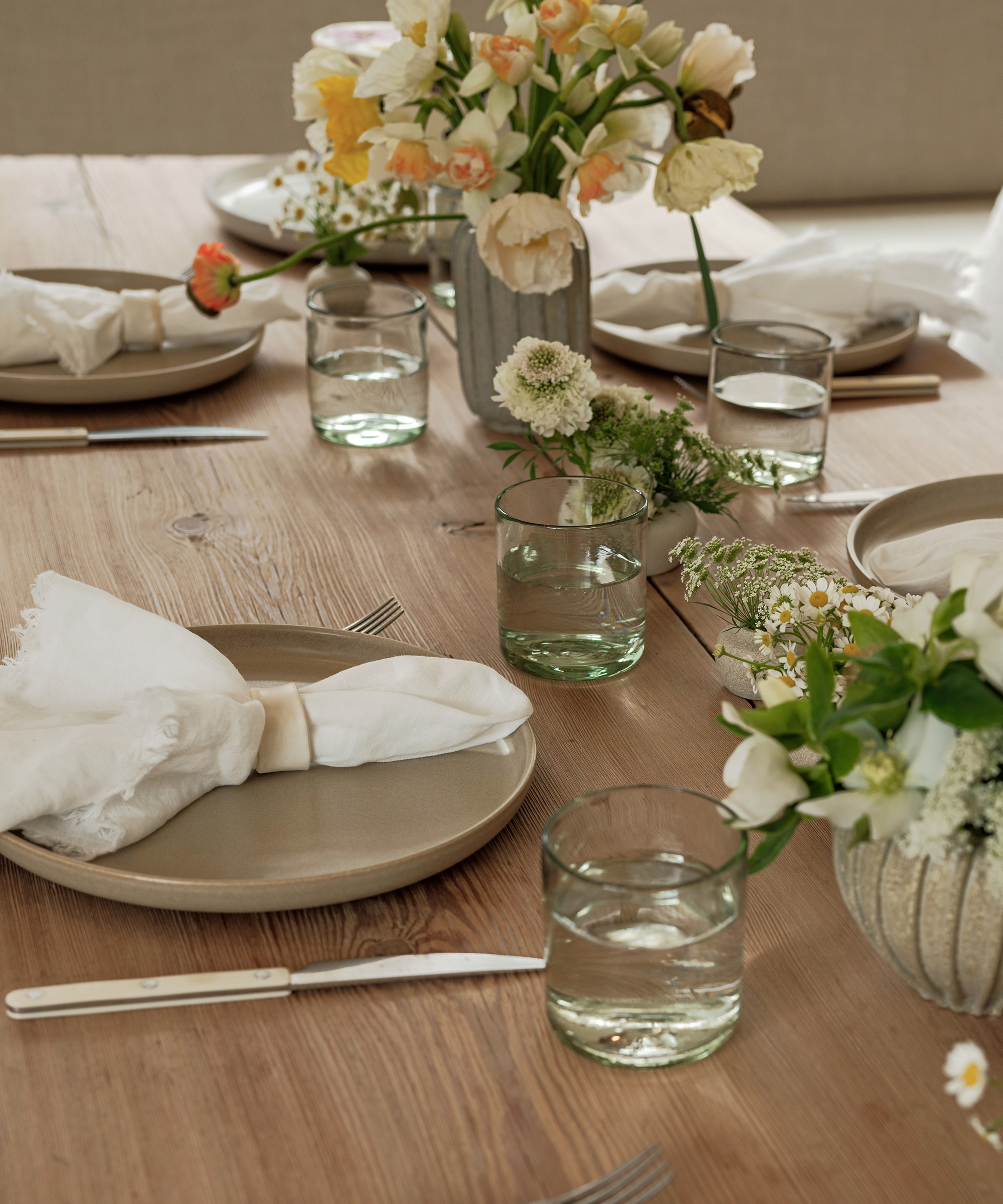 A wooden dining table set with beige plates, white cloth napkins, clear glasses of water, silverware, and vases of assorted white and yellow flowers offers the perfect setting to enjoy delicious cake recipes.