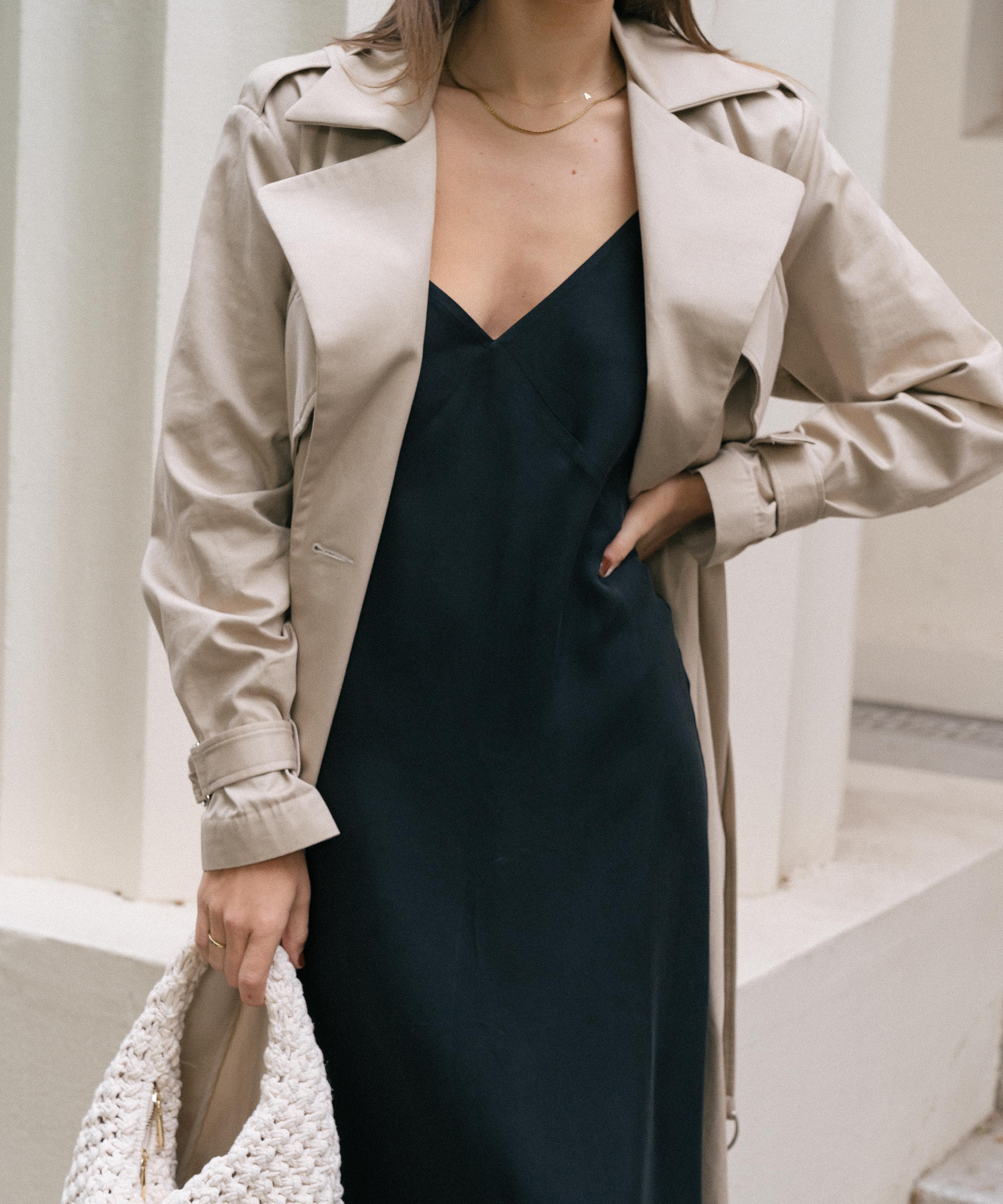 woman in a black slip dress and camel coat holding an ivory purse