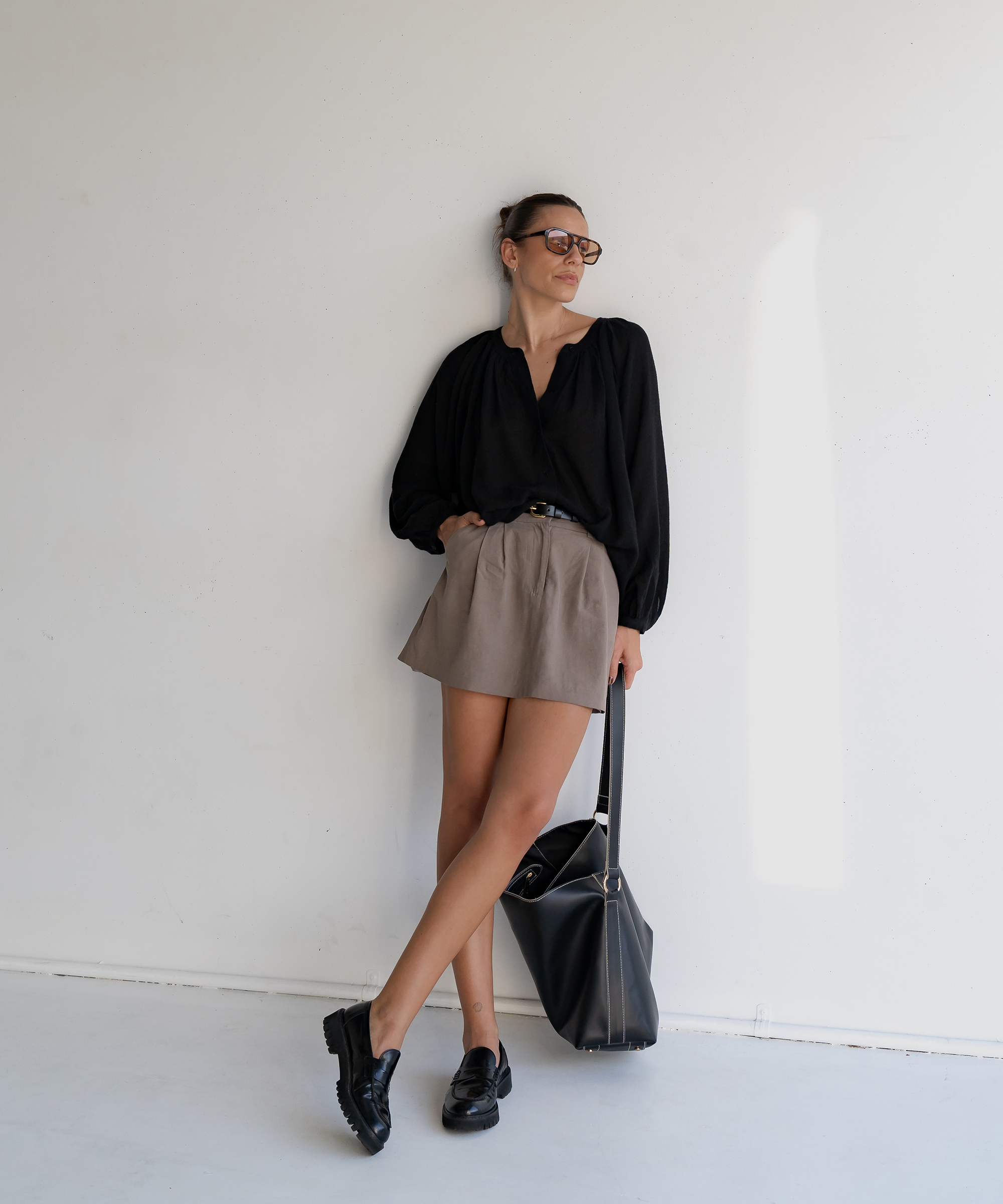 woman standing in a camel mini skirt and black blouse holding a black purse leaning against a wall