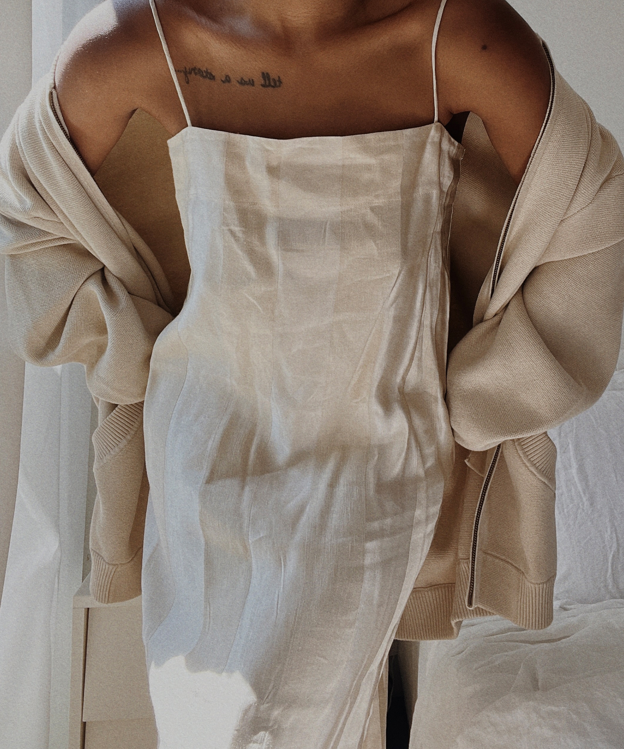 Outfit inspo: A person wearing a light beige slip dress and an oversized beige cardigan, with visible shoulder straps and a chest tattoo, stands in natural light near a window.