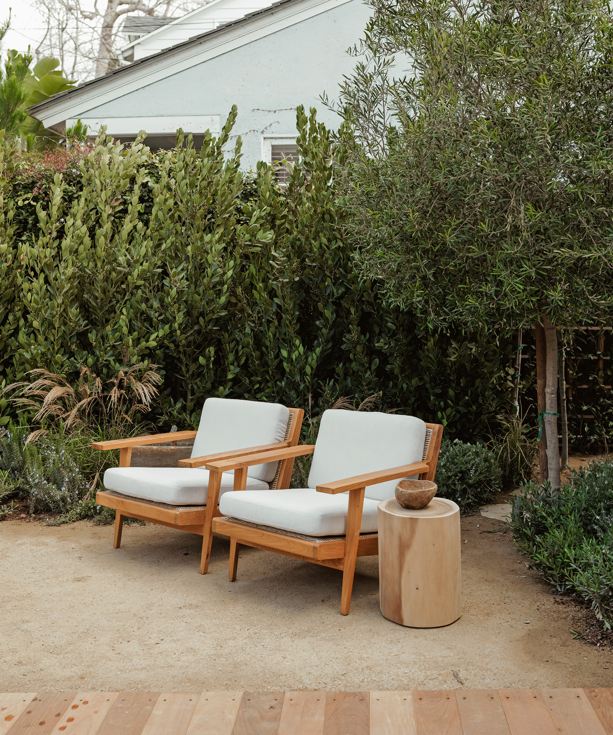 Two wooden lounge chairs with white cushions sit side by side on a patio next to a small round wooden side table, surrounded by lush green plants and trees—perfect inspiration for anyone seeking fresh landscaping ideas for their backyard garden.