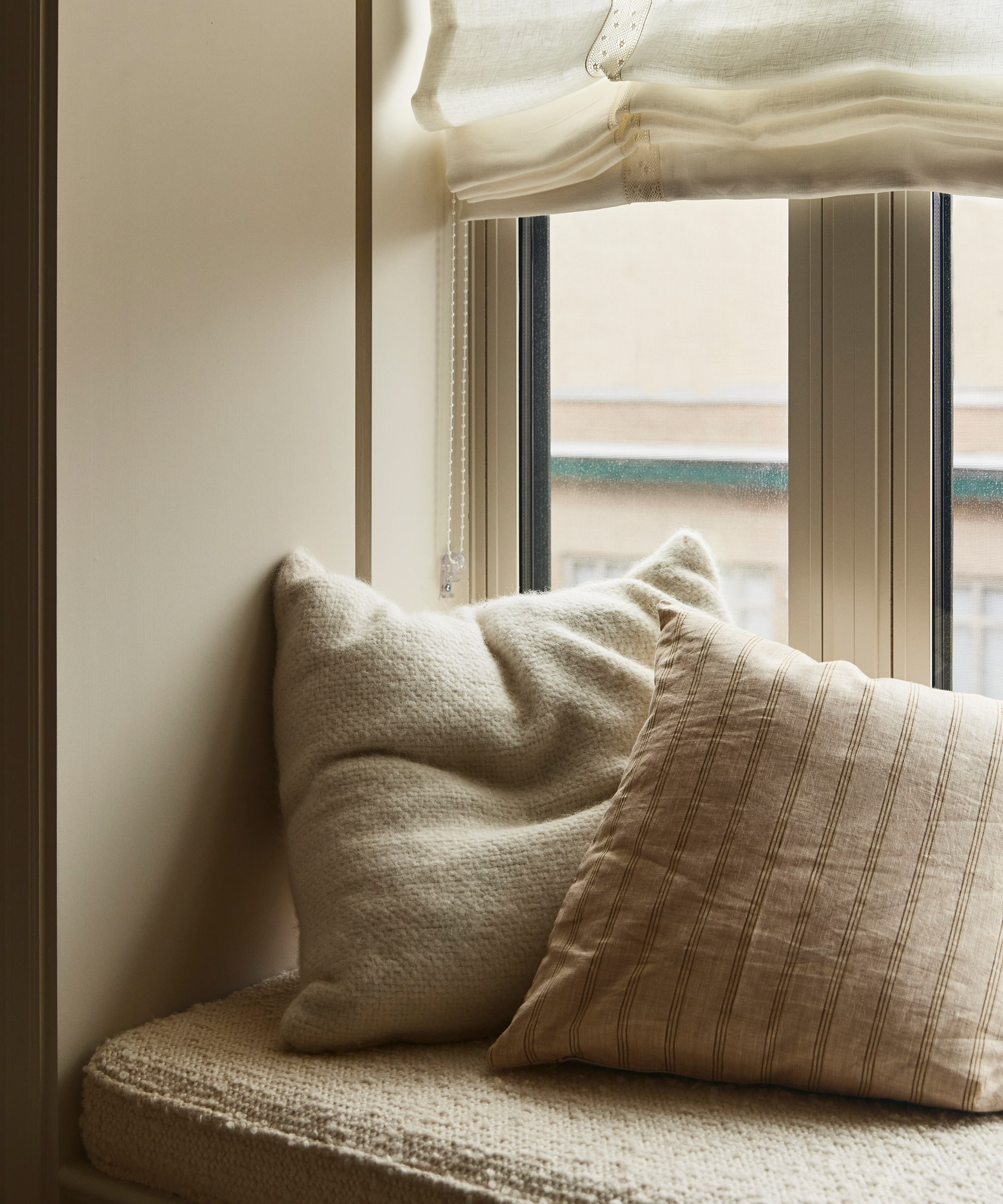 A cozy window seat with a soft, textured cushion and two pillows—one cream and fluffy, the other beige with stripes—sits beside a window with a partially raised white roman shade in a charming New York City apartment.