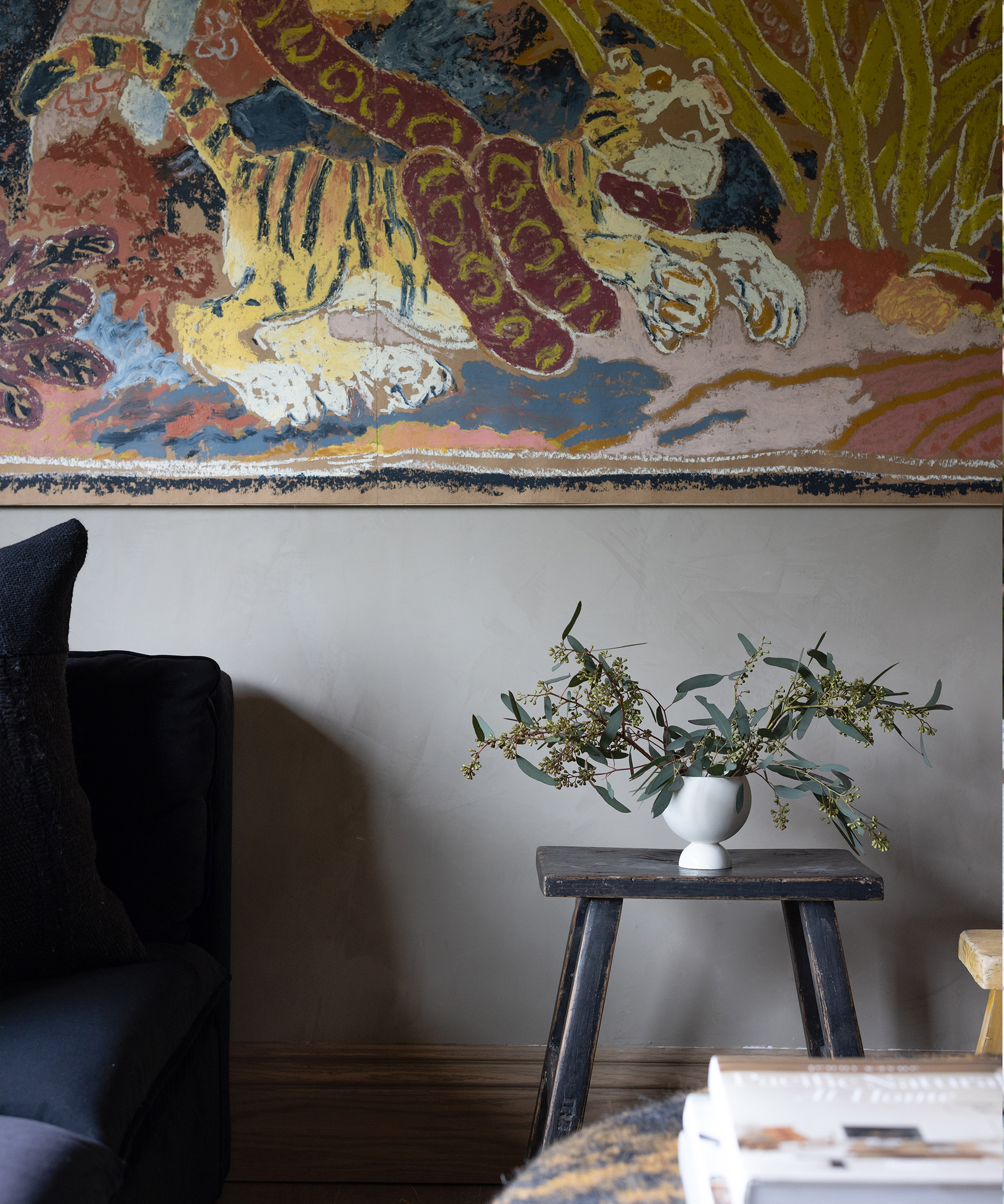 A dark wooden stool with a white vase holding green branches sits beside a dark sofa. Above, a colorful wall tapestry features a tiger and foliage. Part of a table and book are visible in the foreground.