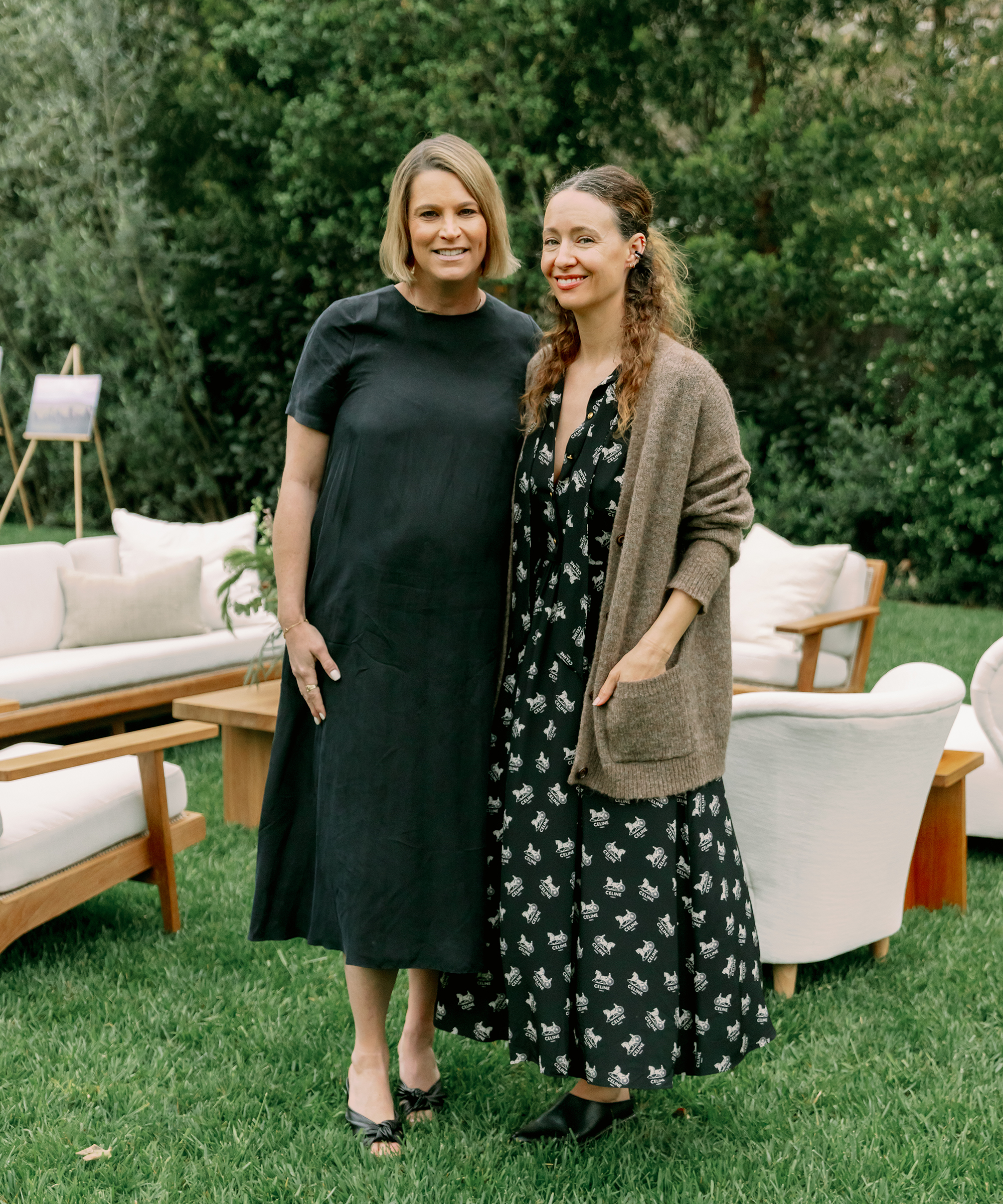 two women smiling and posing in the grass wearing a black maxi dress
