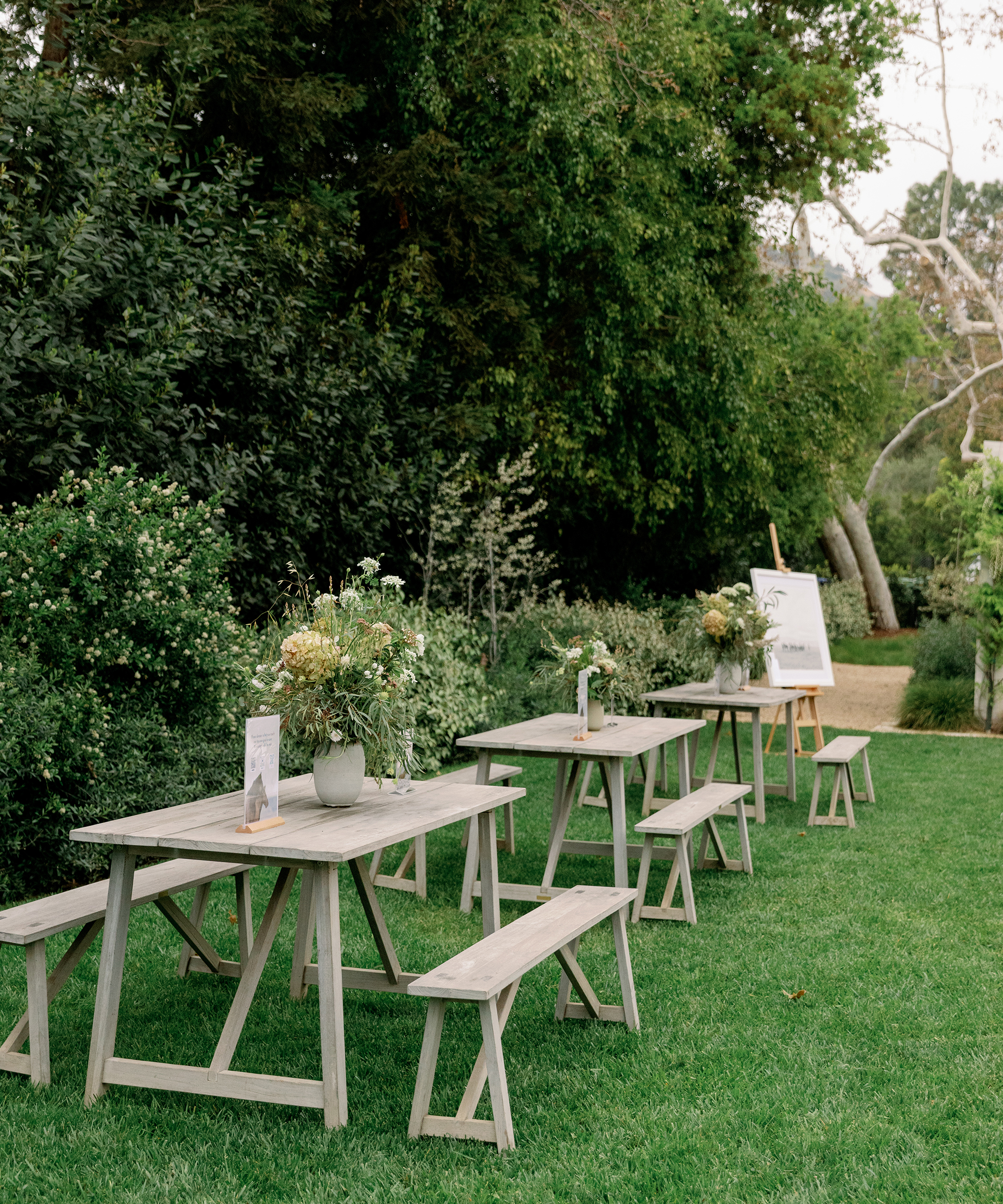 Three rustic wooden picnic tables with benches are arranged on a grassy lawn, each decorated with flower arrangements. Lush green trees and shrubs surround the area, evoking the spirit of the American wild horse, with a white signboard in the background.