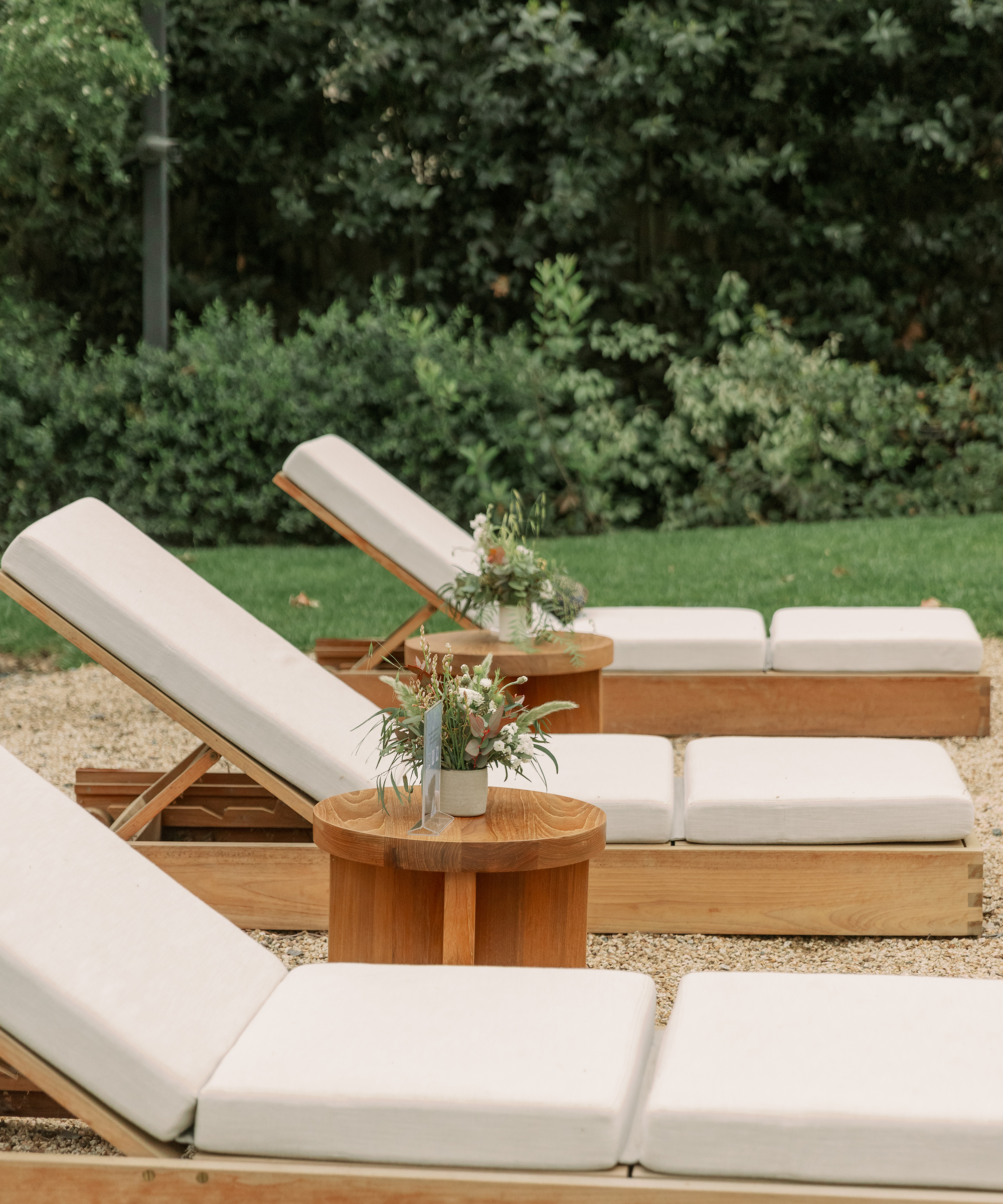 Three wooden lounge chairs with white cushions are lined up outdoors on a gravel surface. Small round wooden tables with potted floral arrangements sit between the chairs, while lush green bushes evoke the untamed beauty of the American wild horse in the background.
