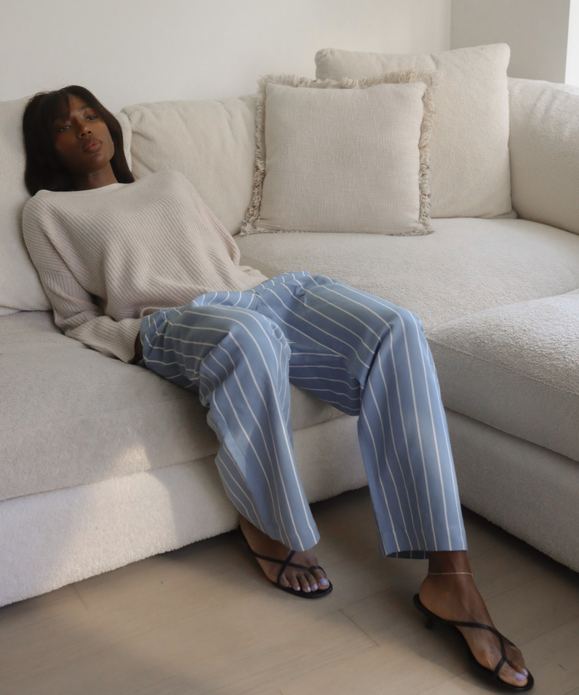 A woman in a beige sweater and light blue striped pants sits relaxed on a cream-colored sectional sofa, with neutral decor and soft natural light in the room.