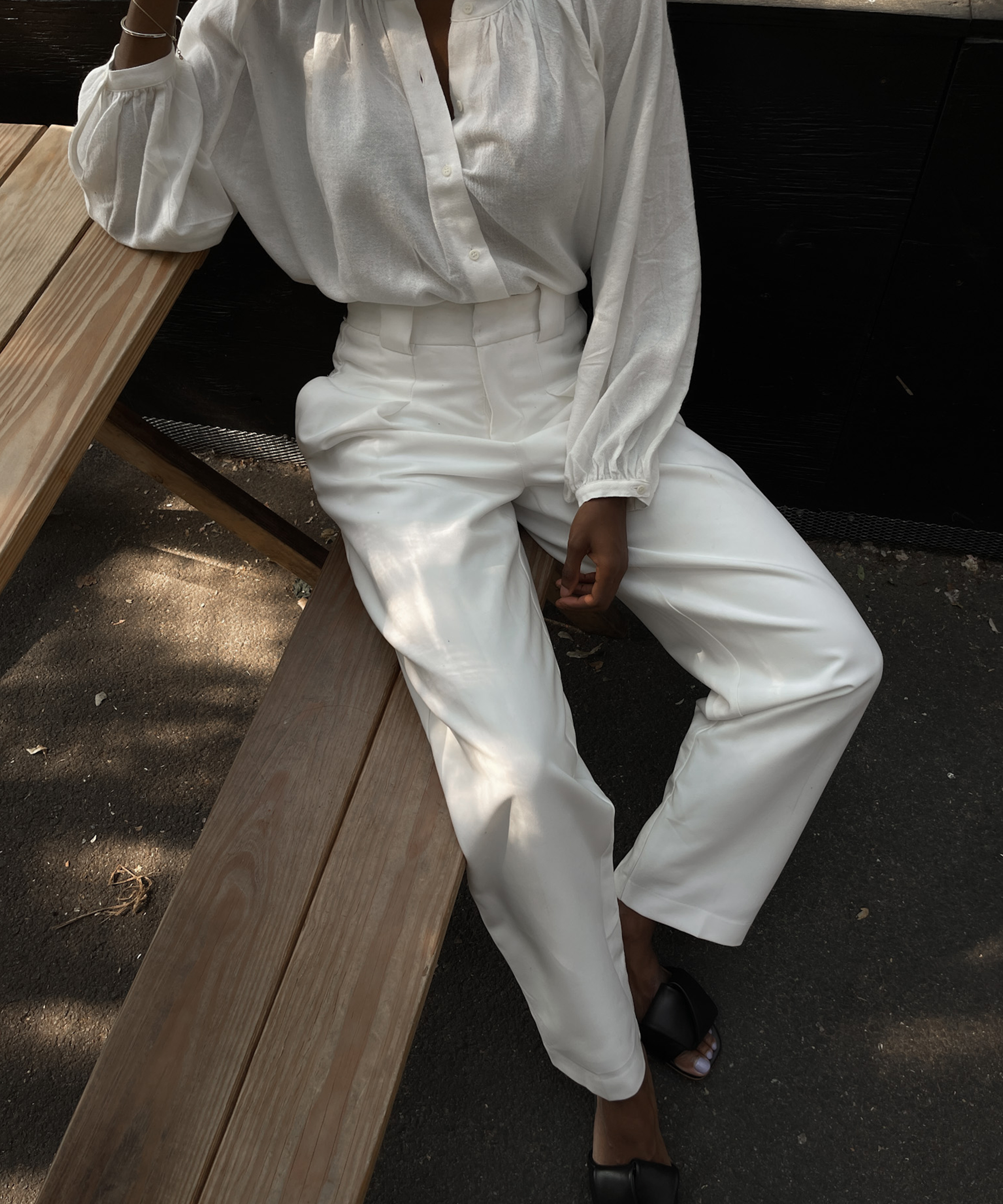 woman sitting on a wooden bench in white trousers and a white blouse with black slides