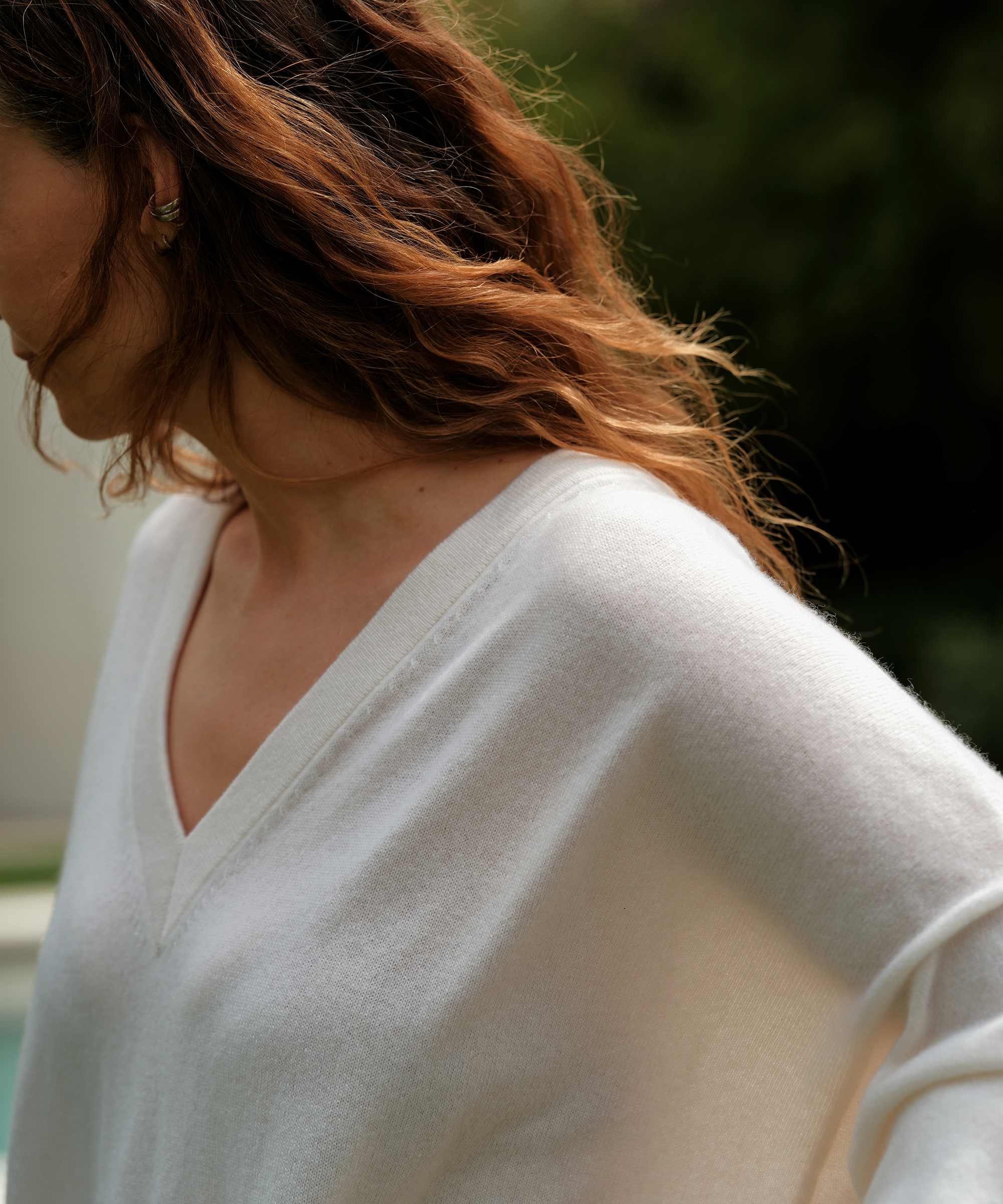 A woman with wavy, auburn hair wears a white V-neck sweater—an effortless nod to summer style. She stands outdoors near greenery, facing slightly away from the camera, her left arm bent and her face mostly out of frame.
