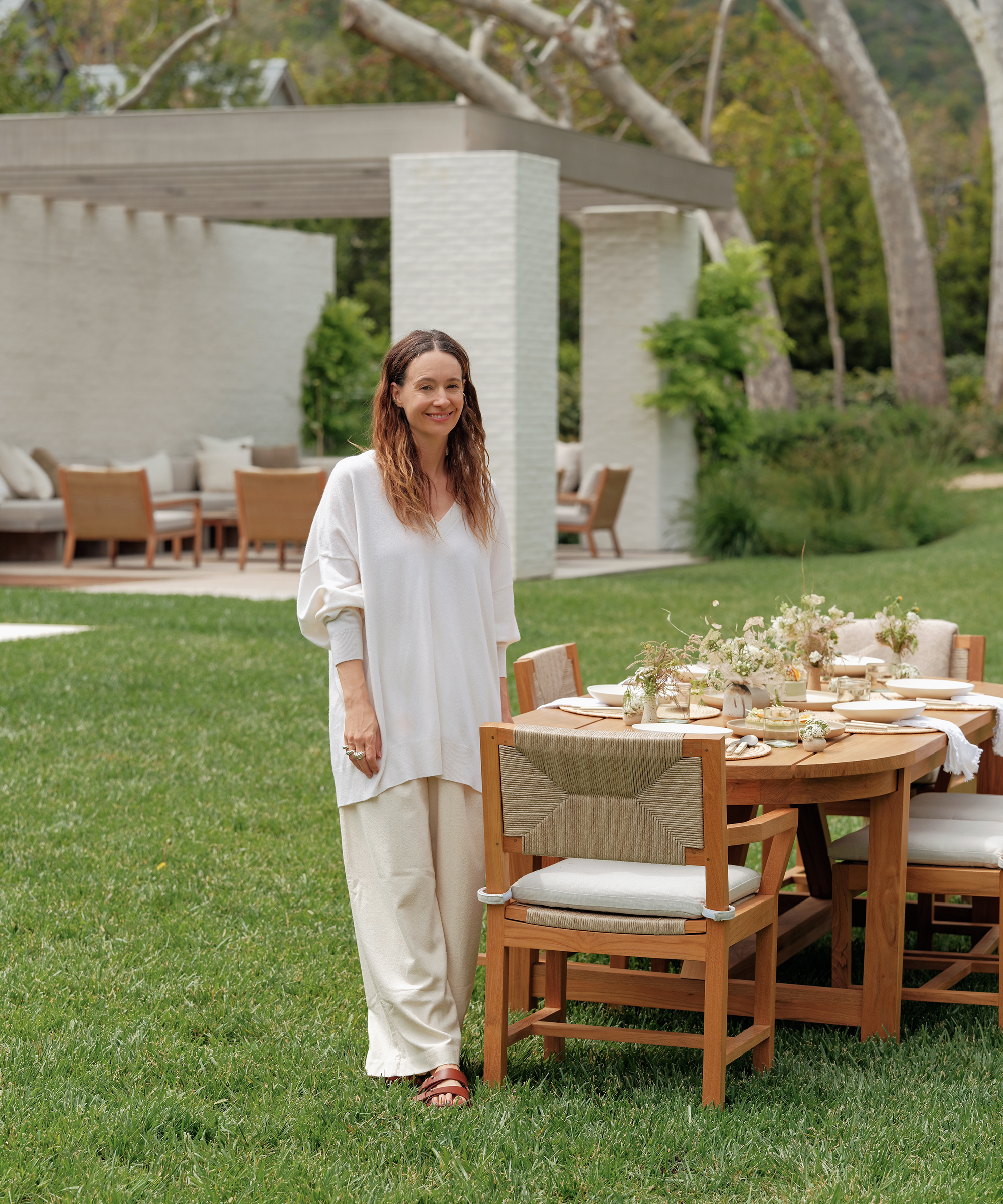 A woman in loose, light-colored clothing stands smiling next to a wooden outdoor dining table set with plates, glasses, a vegan cake, and floral arrangements on a green lawn, with a patio and trees in the background.