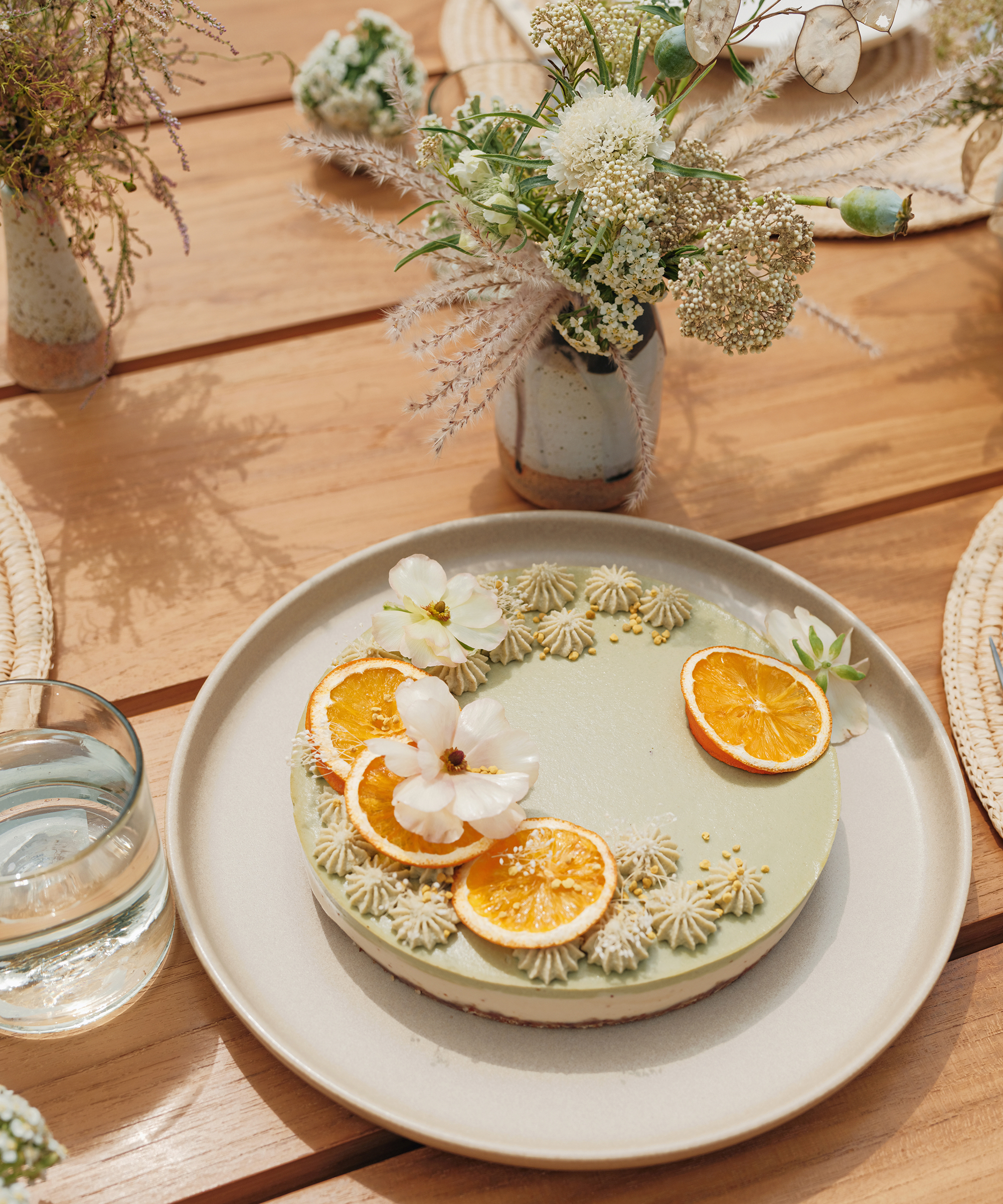 A round, light green vegan cake topped with dried orange slices and white flowers sits on a beige plate on a wooden table, surrounded by glassware, woven placemats, and small vases of white wildflowers.