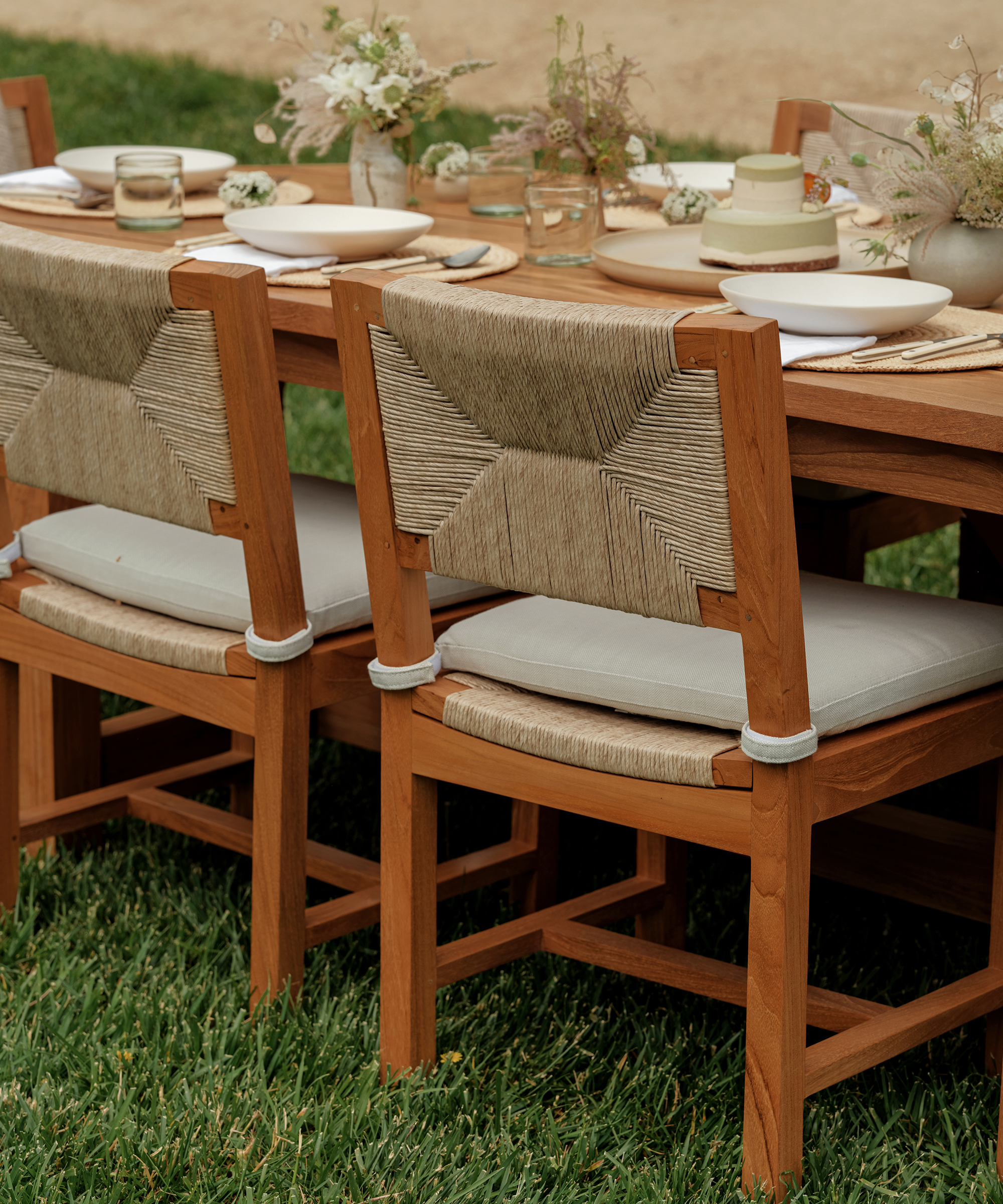 A close-up of a wooden outdoor dining table set for a meal, with two wooden chairs featuring woven backs and light cushions, on green grass. The table is decorated with plates, glasses, small floral arrangements, and a tempting vegan cake.
