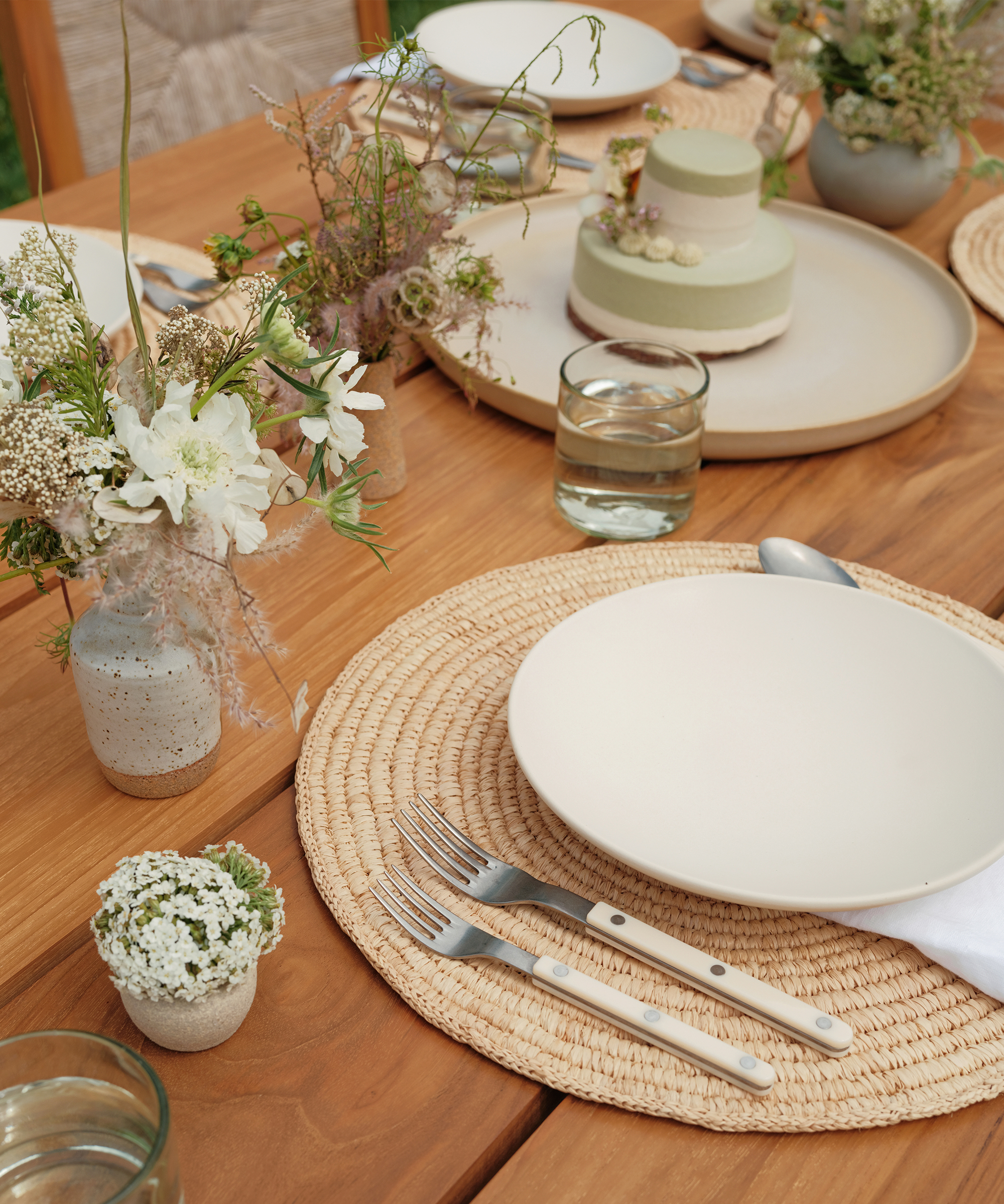 a wood outdoor dining table with a braided placemat, ivory sierra dinner plate, ivory bistro vintage cutlery and green and white florals