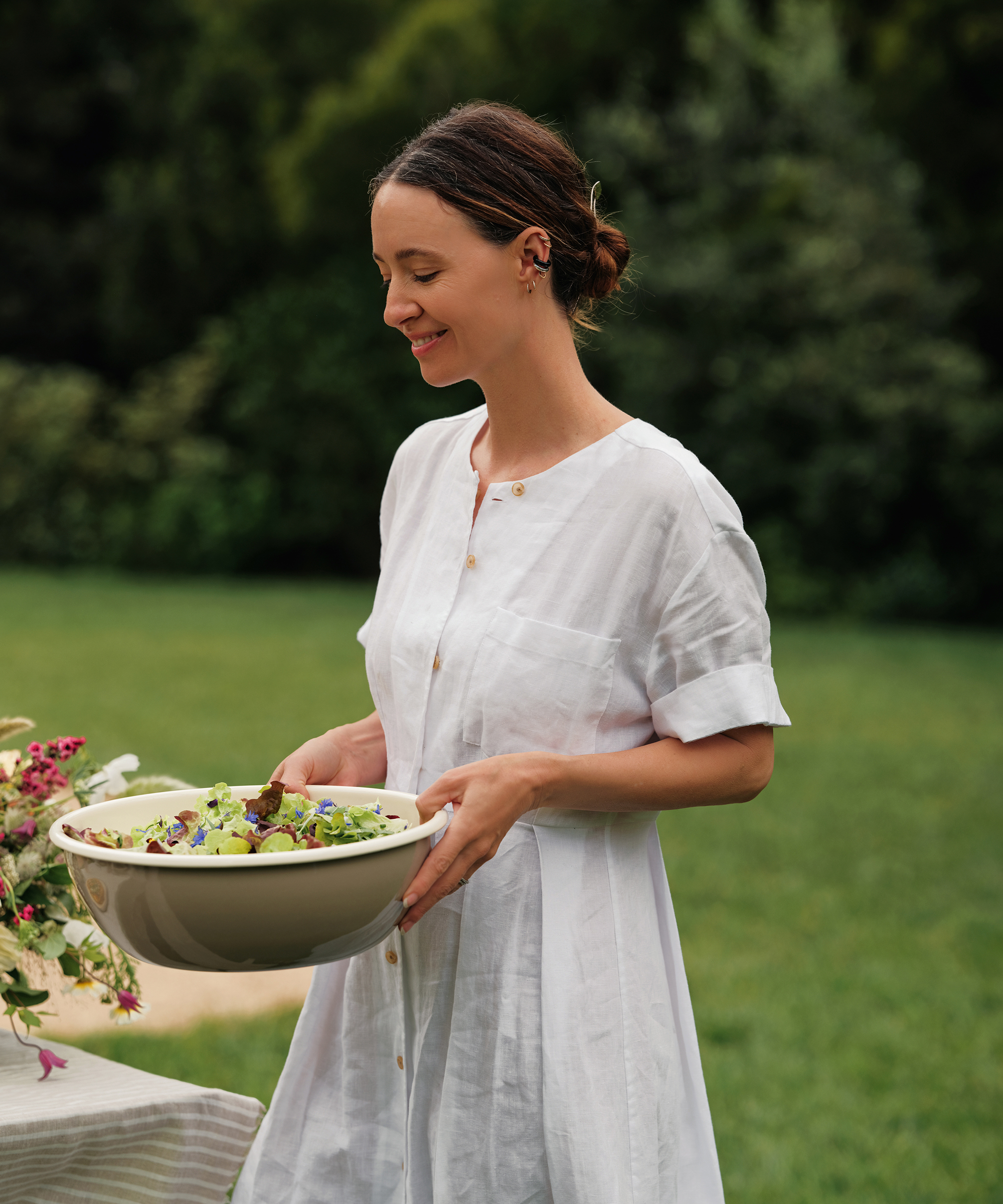 jenni kayne in a white linen day dress holding a taupe and cream outdoor serving bowl with a salad