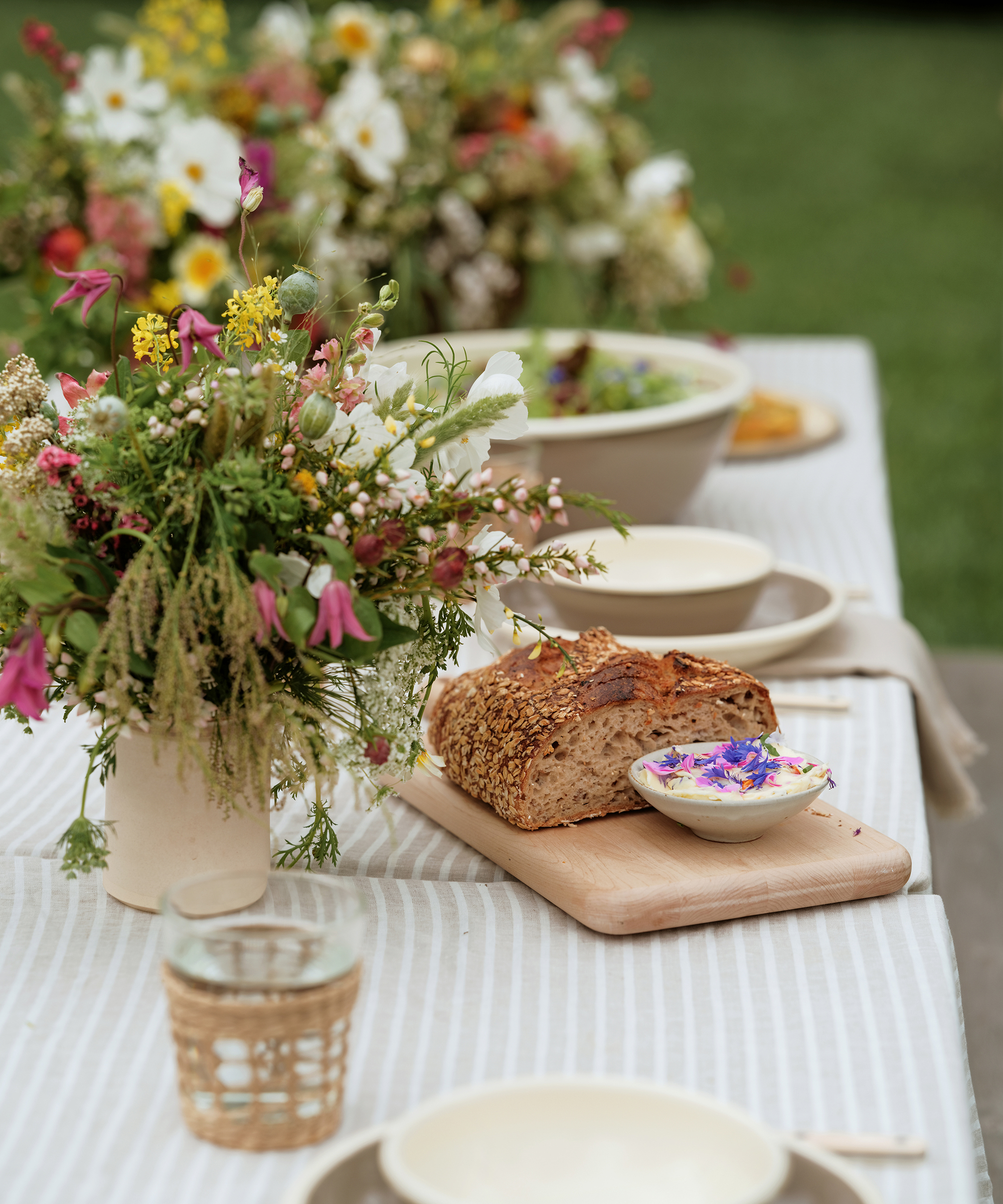 A rustic outdoor table is set with wildflower bouquets, a loaf of bread on a wooden board, herb butter with edible flowers, and glasses on a striped tablecloth—perfect inspiration for easy summer recipes.
