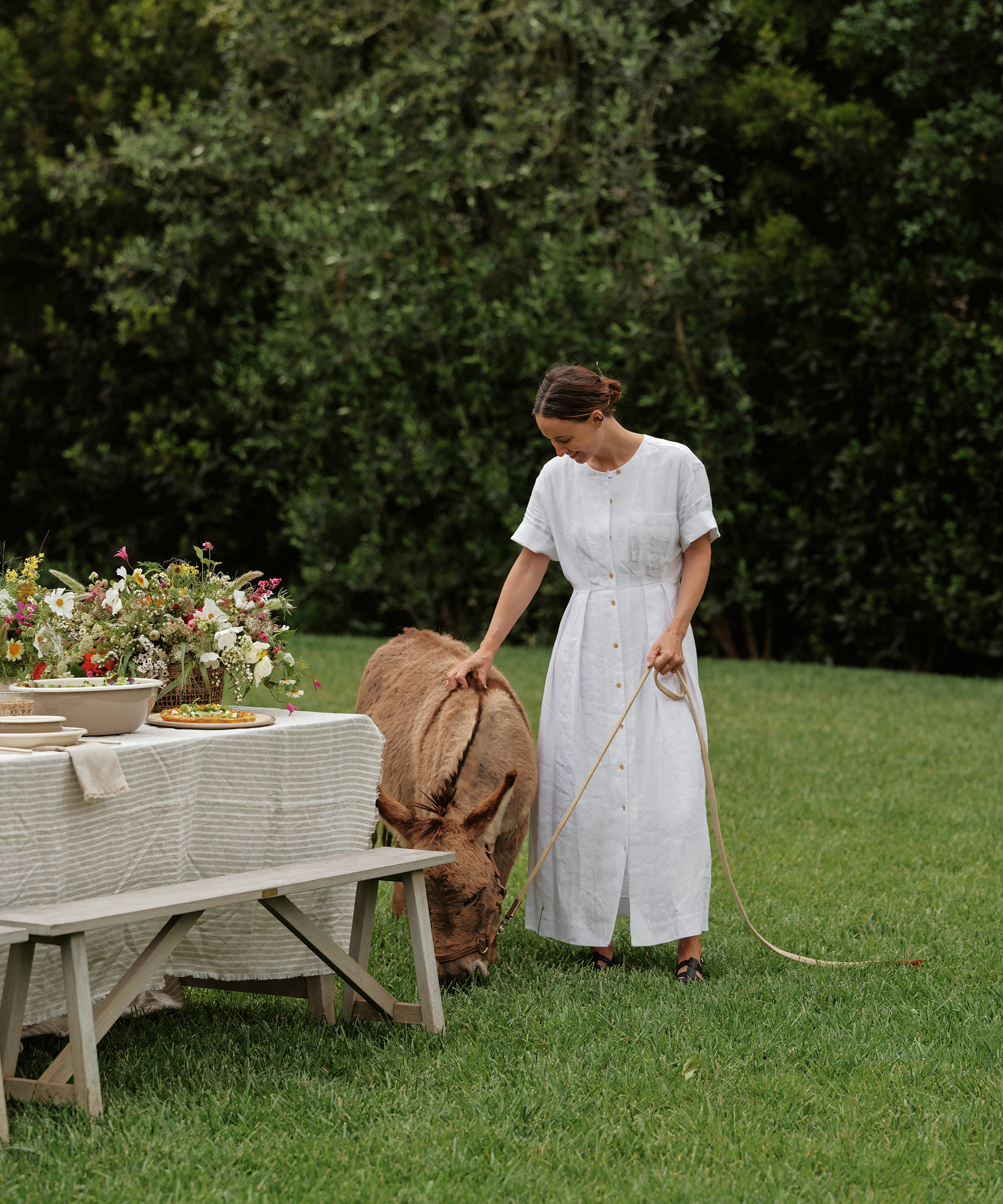 jenni kayne wearing a white linen day dress and black sandals walking her donkey in the grass next to a picnic table with striped linens
