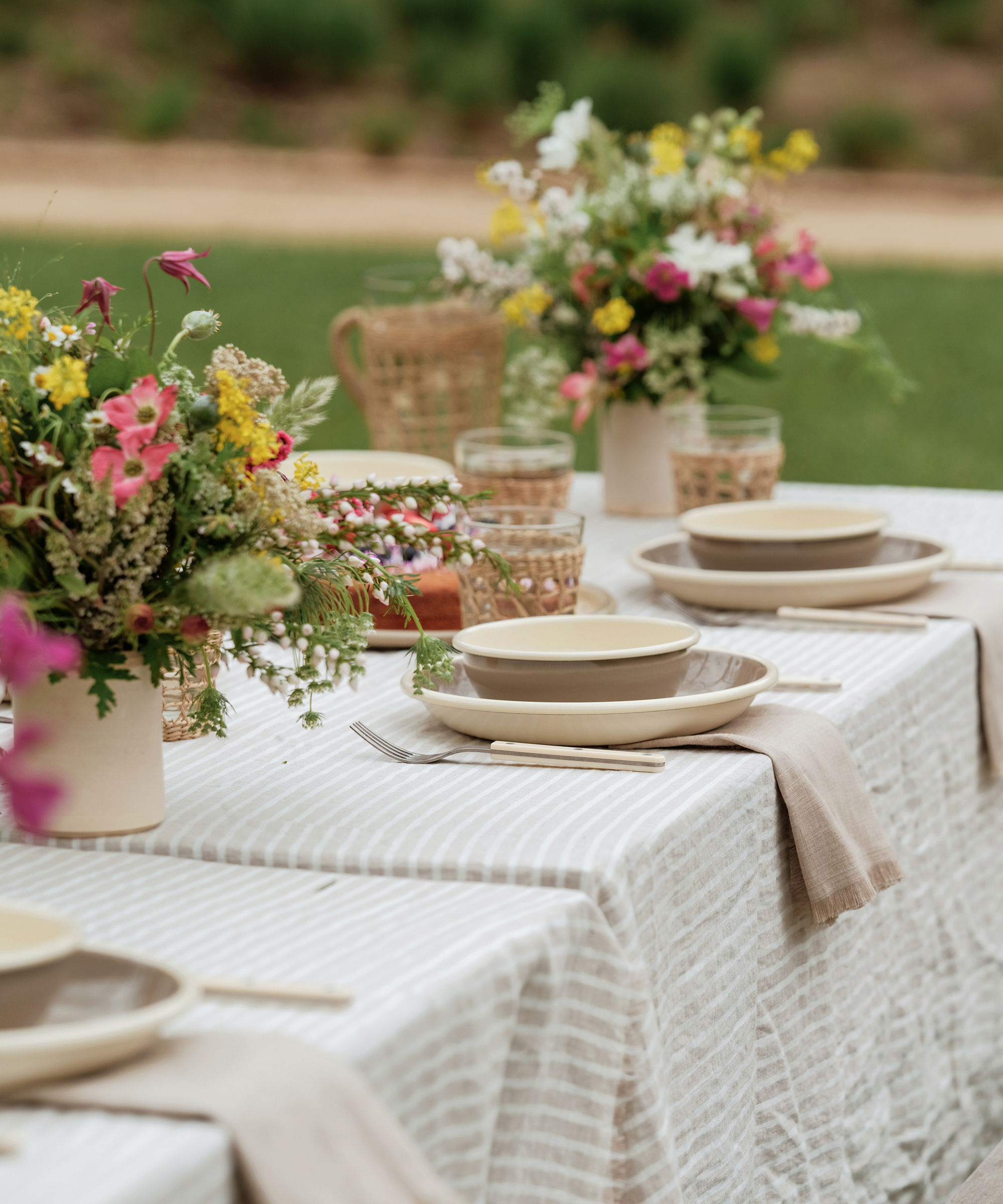 a striped linen table cloth taupe and cream outdoor dinner plates and bowls, colorful flowers and rattan glassware on an outdoor picnic table
