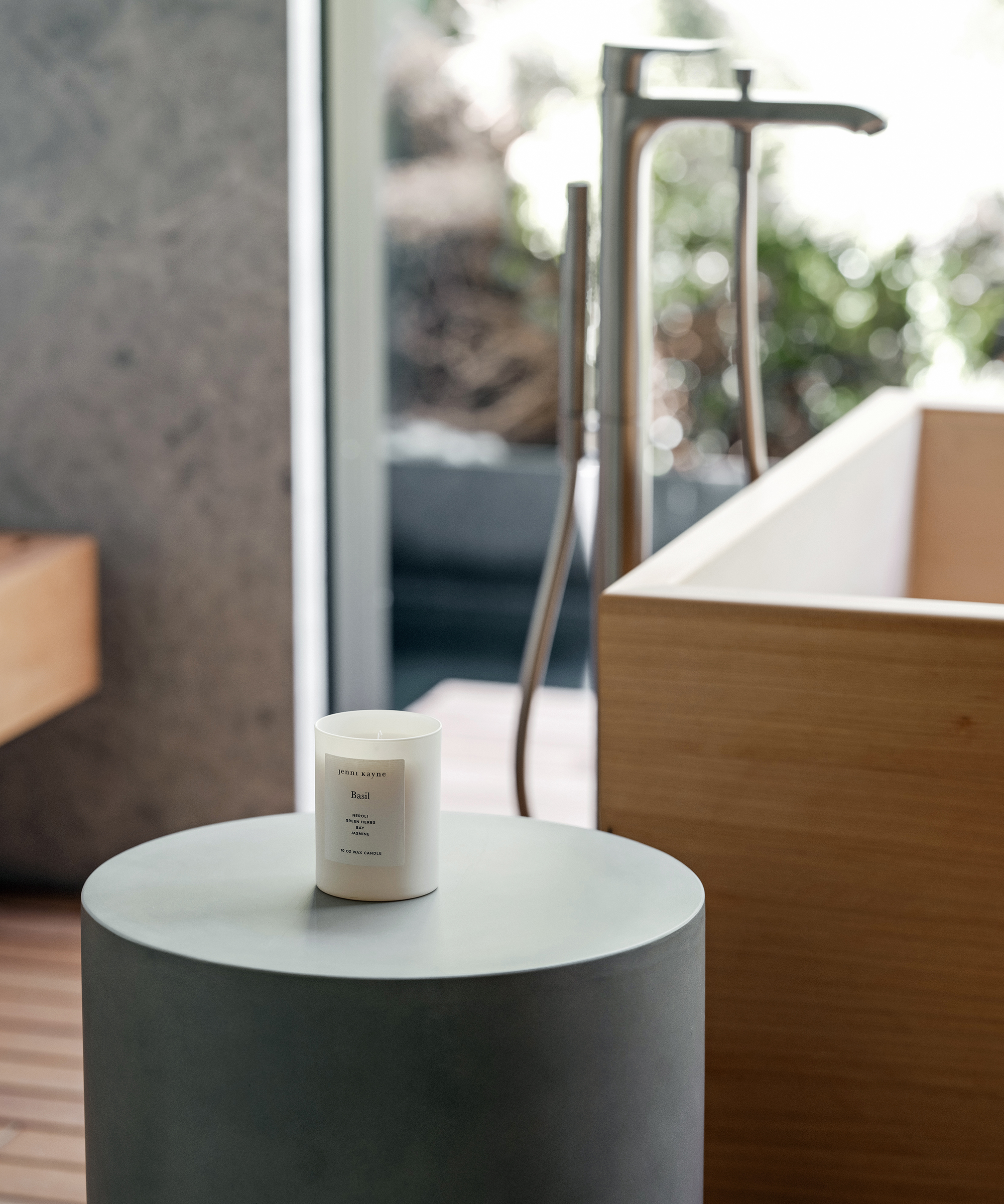 A white candle in a glass jar sits on a gray round table beside a wooden bathtub and modern chrome faucet in a bright, Palm Springs-inspired bathroom with large windows and lush greenery outside.