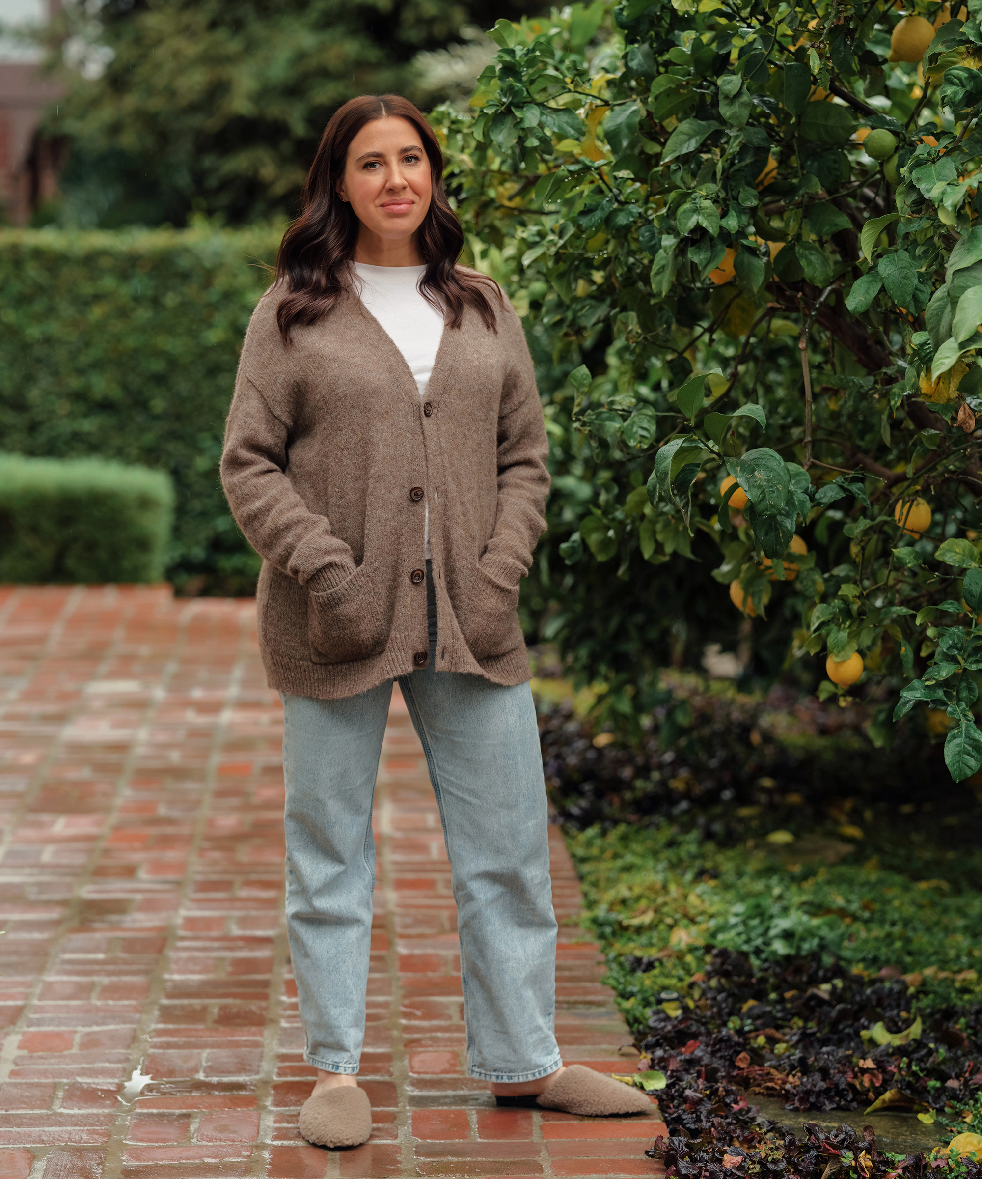 aliza standing outside on a brick path next to a lemon tree in jeans and a brown cardigan with a white tee and shearling mules