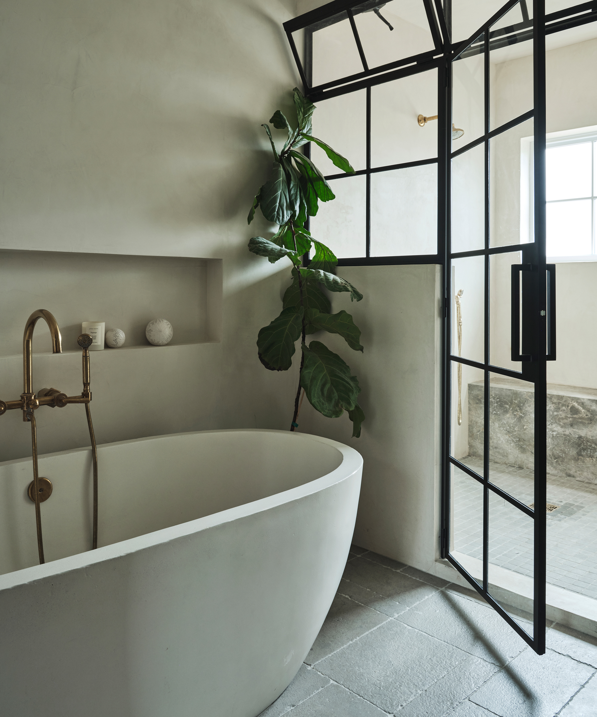 Minimalist bathroom in a Montecito home featuring a white freestanding tub, brass fixtures, a potted green plant, gray stone floors, and a glass black-framed shower door letting in natural light.