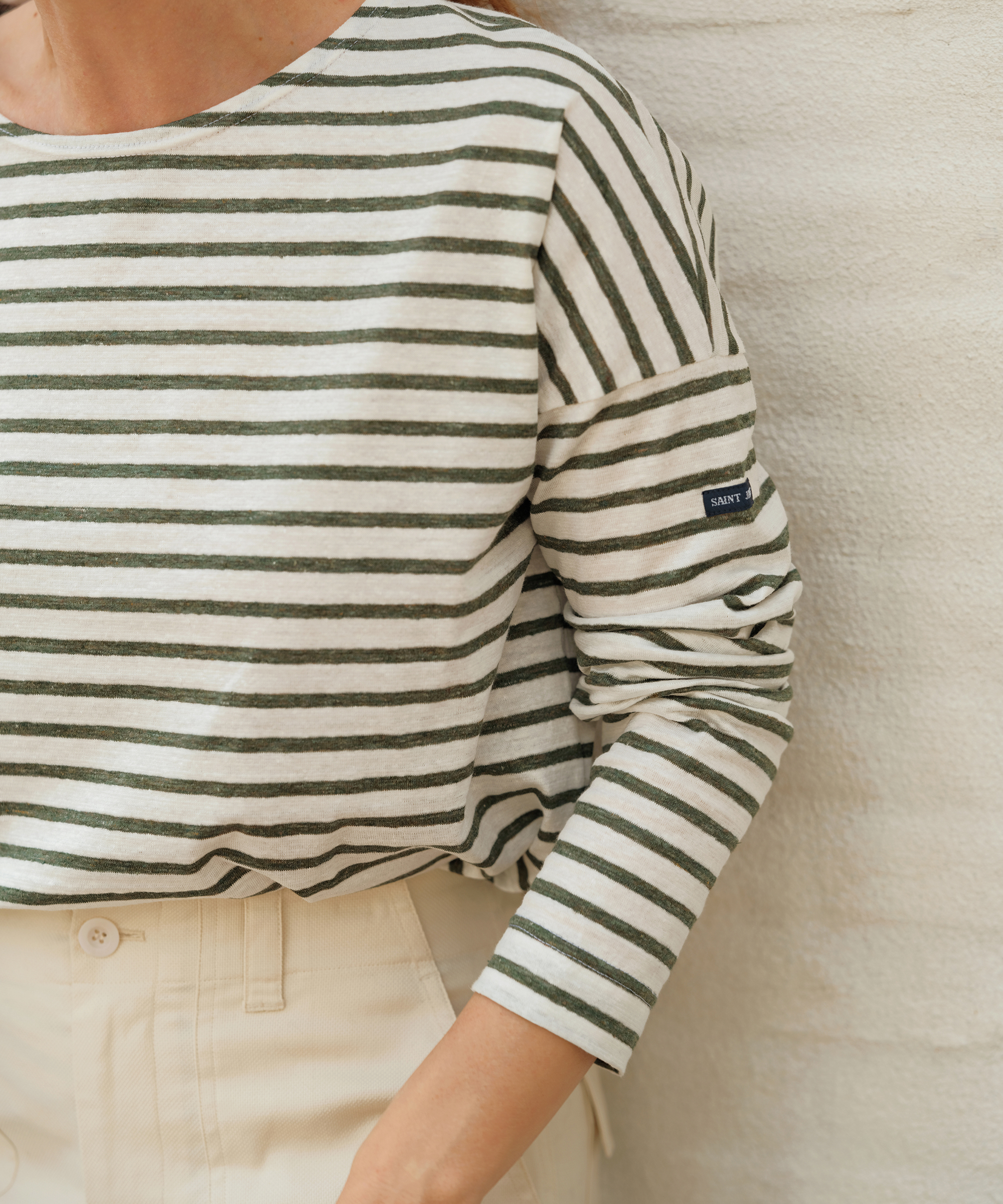 jenni kayne wearing the a long sleeve striped tee with olive stripes leaning against an ivory brick wall