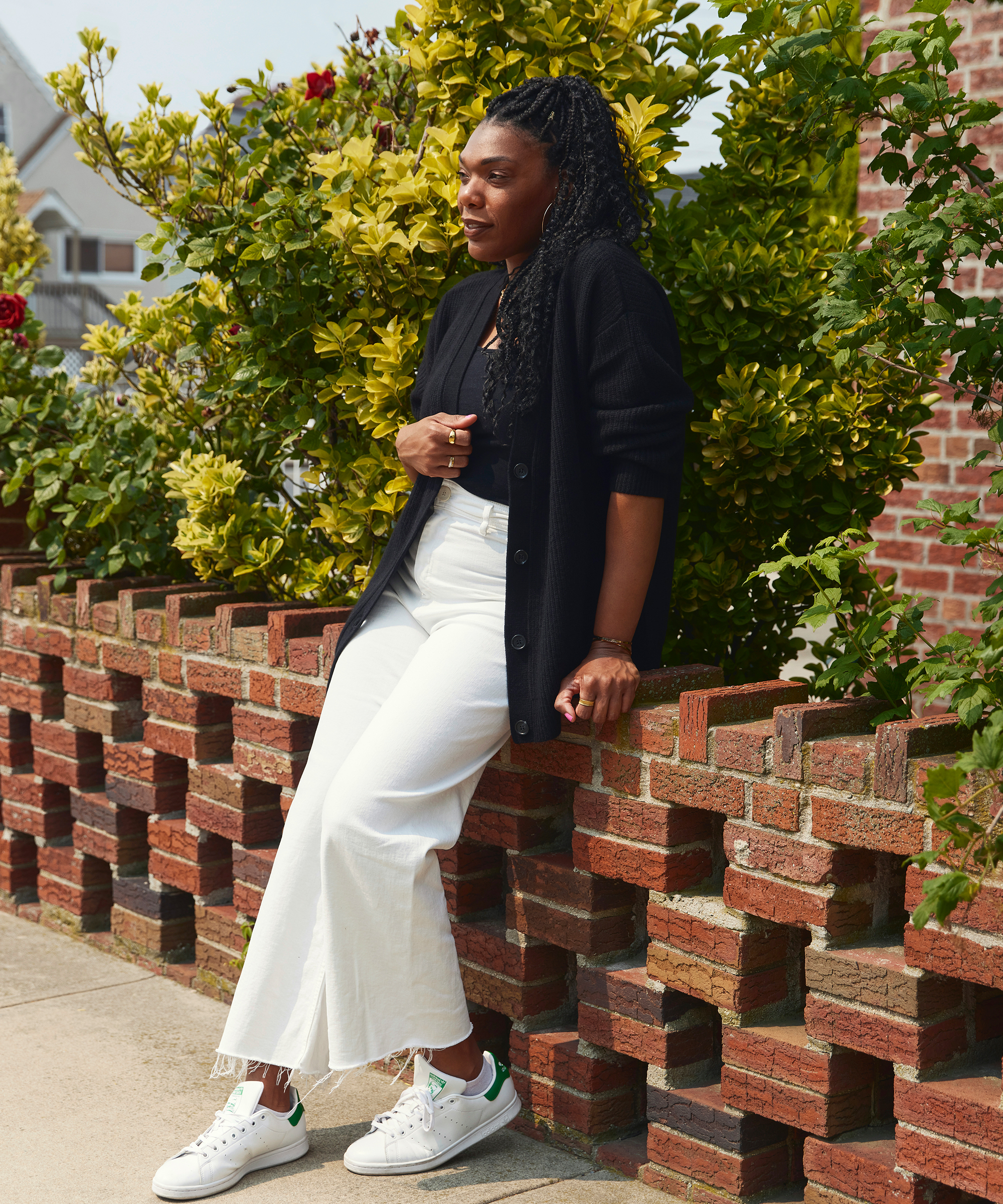 A wellness coach with long braids, dressed in a black top and cardigan with white wide-leg pants and white sneakers, leans on a brick wall outdoors next to green and yellow bushes.