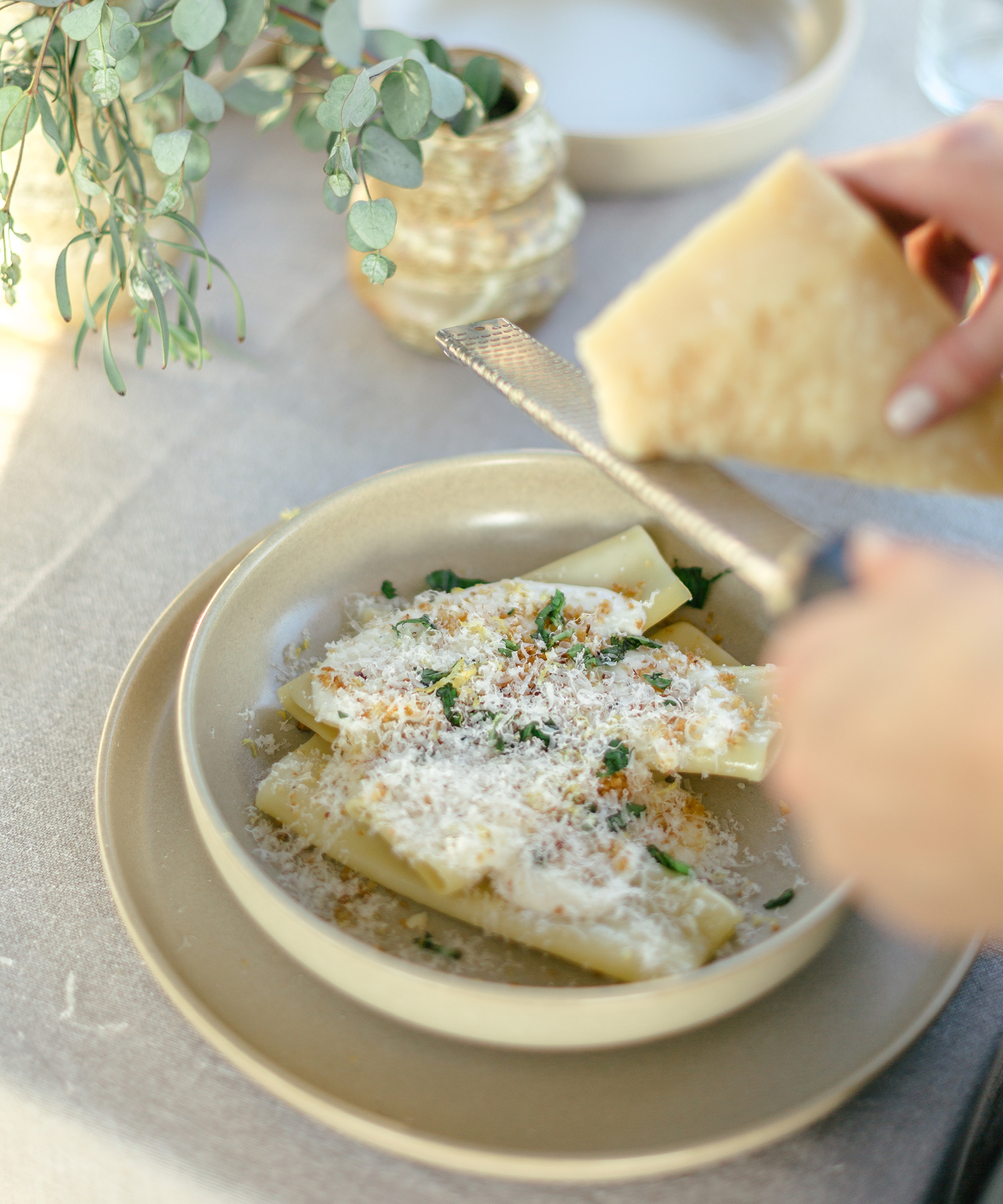 grating parmesan into a taupe bowl of pasta on a linen placemat