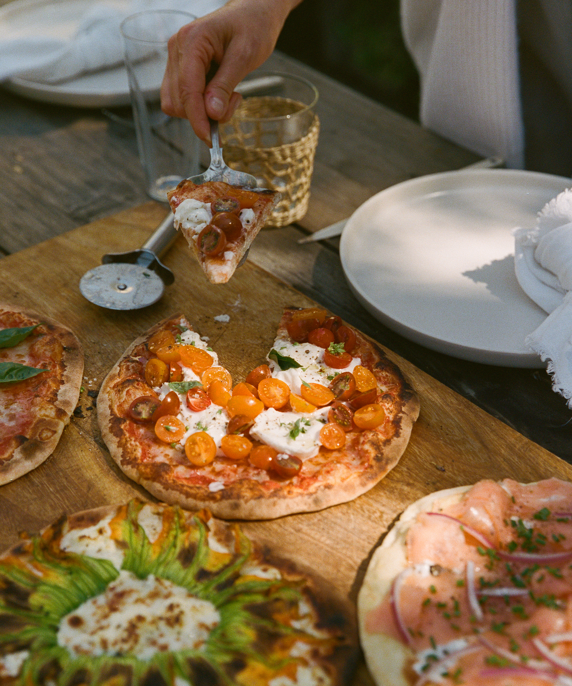 A hand lifts a slice of pizza topped with cherry tomatoes, cheese, and basil from a wooden board with several pizzas—ideal for a relaxed Labor Day gathering. Empty plates, a glass of water, and a pizza cutter rest on the rustic table.